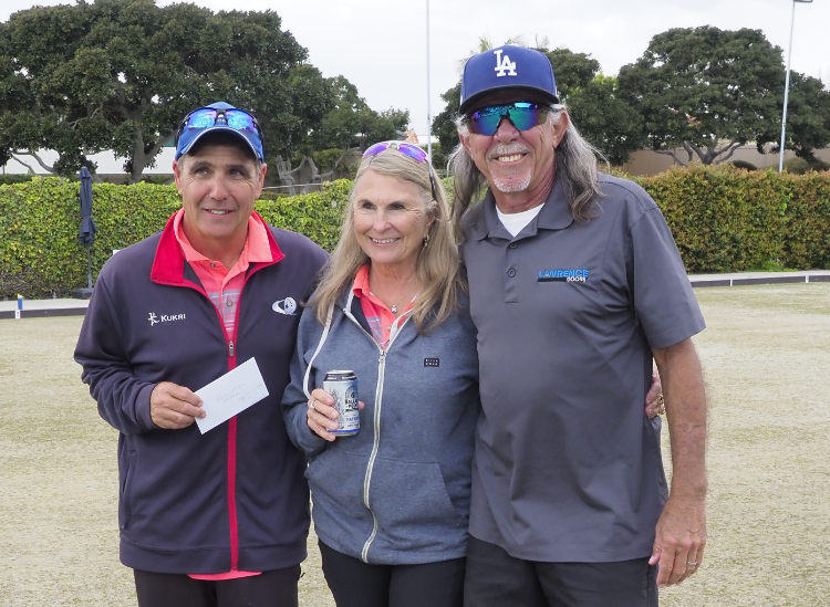 Three people standing outdoors on a lawn, smiling at the camera, with trees and a hedge in the background. The person on the left is holding an envelope, the woman in the middle is holding a soda can, and they are all dressed in casual outdoor attire, including hats and sunglasses.