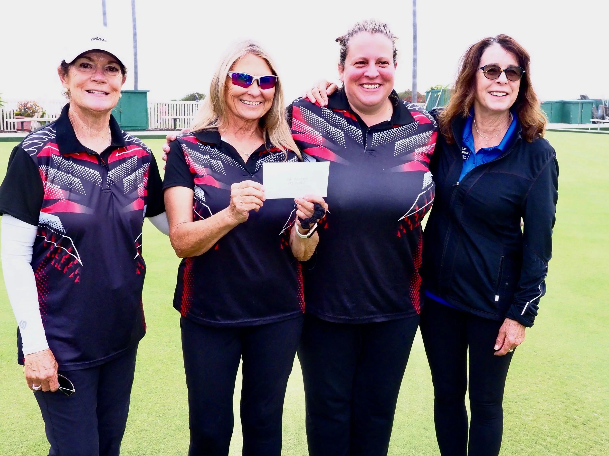 Four women standing together on a golf course, smiling, with one woman holding a check.