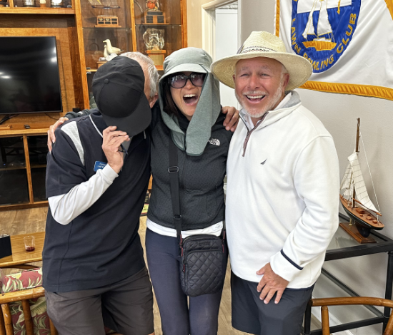 Three people in a cheerful group hug, smiling, with a woman wearing sunglasses and a sun hat, and two men, one wearing a cap and the other a wide-brimmed hat, inside a room with nautical decor.