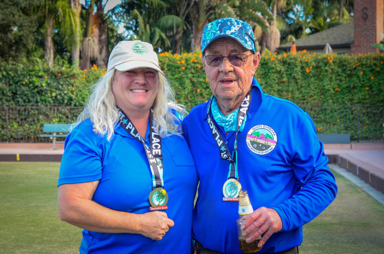 Two smiling older adults standing outdoors on a lawn, wearing blue shirts and medals, with lush greenery and trees in the background.