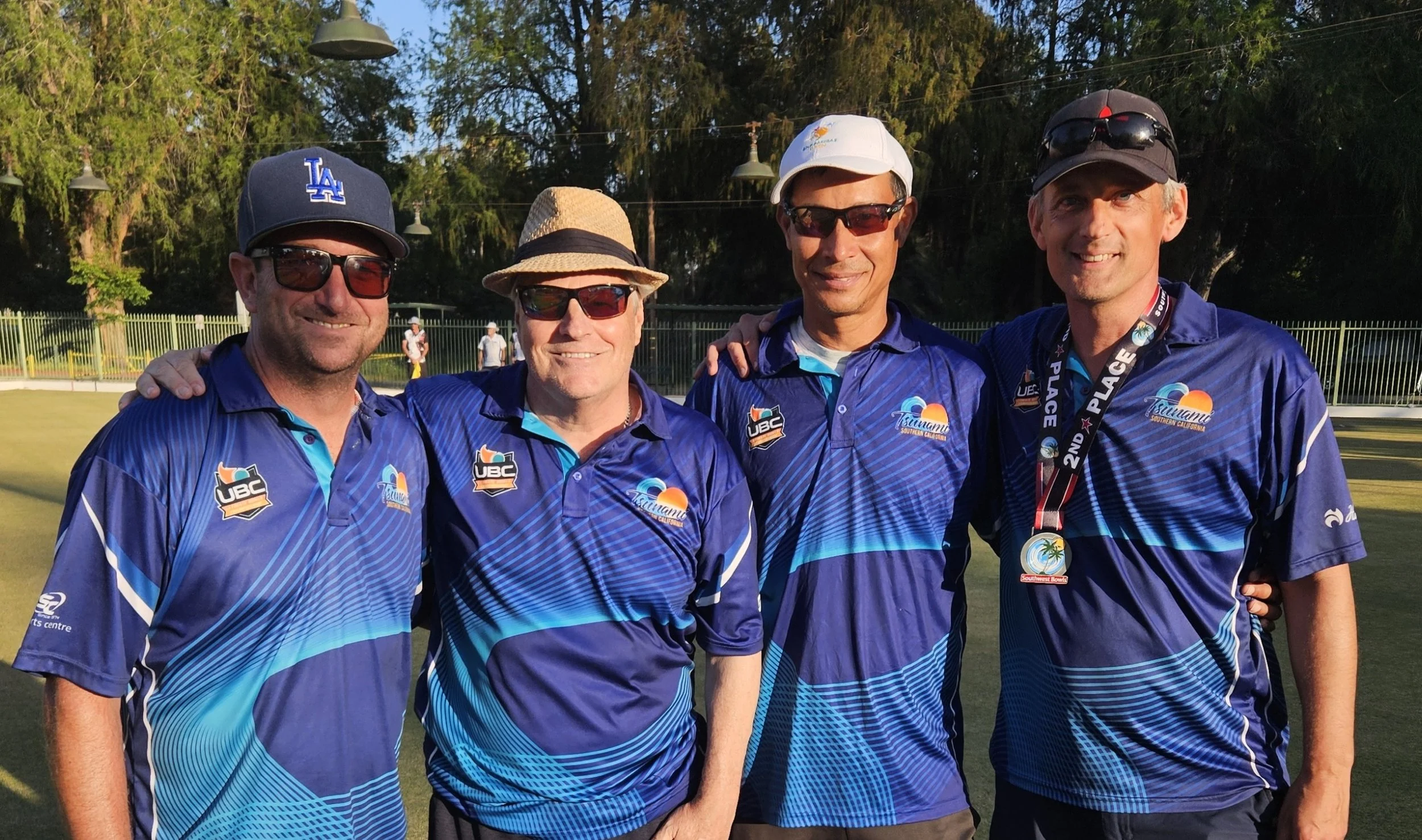 Four men wearing blue sports jerseys with logos, sunglasses, and hats, standing outdoors on a green field with trees in the background, smiling and posing for a photo.
