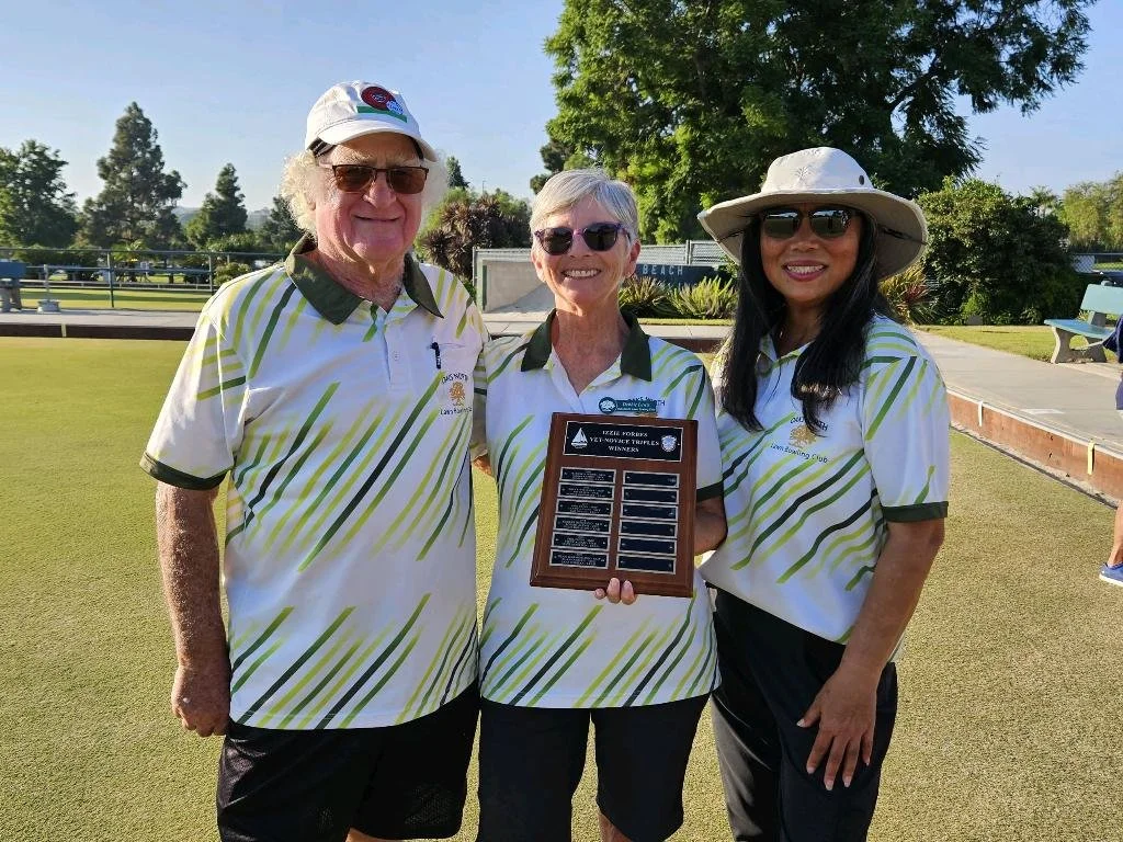 Three people standing on a lawn, smiling, holding a plaque, with trees and a fence in the background. They are wearing matching sports jerseys with green and white stripes, and sunglasses. One person is wearing a wide-brimmed hat.