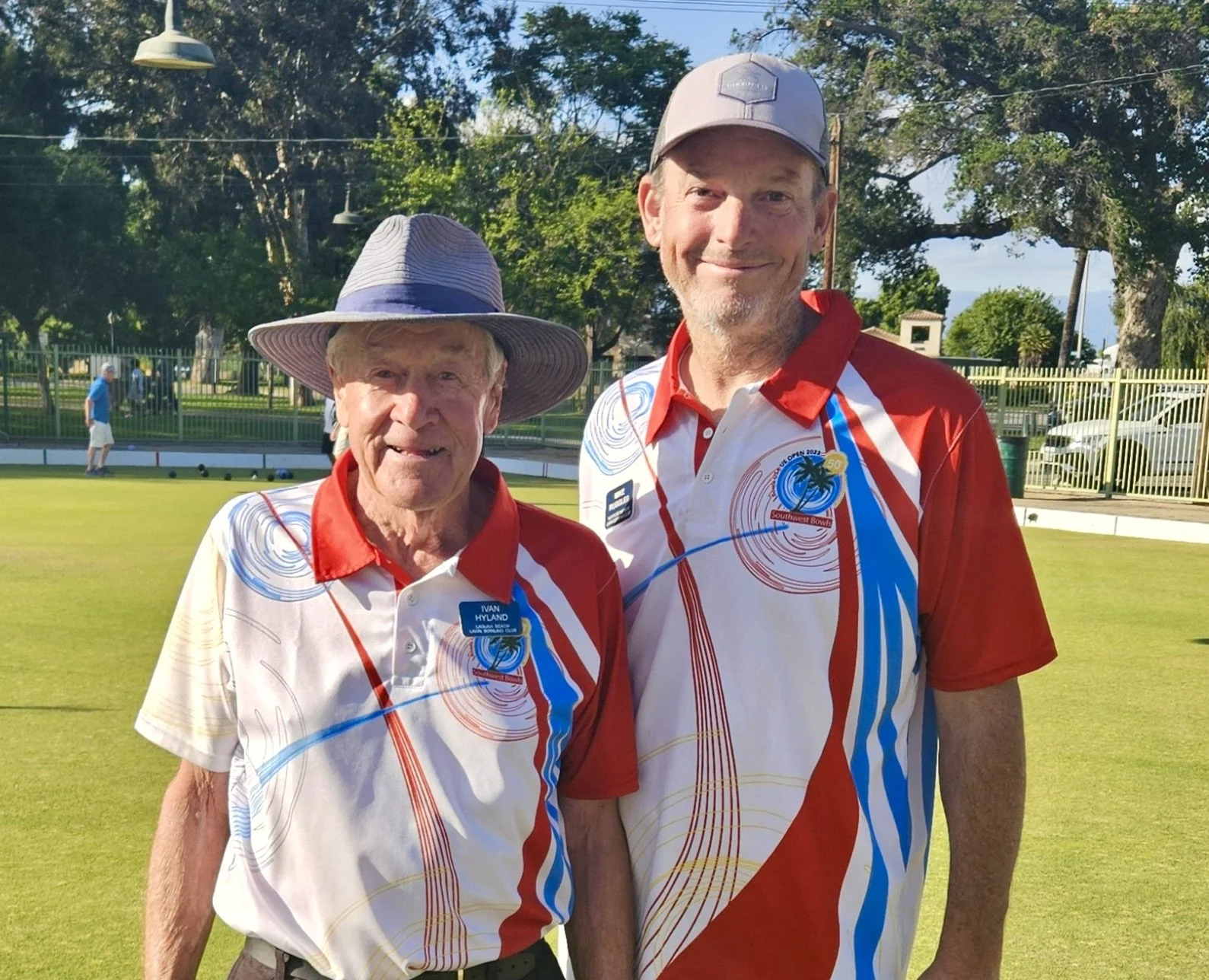 Two men standing on a lawn, wearing matching sports shirts with red, white, blue, and gold designs, at a lawn bowls game. The older man on the left wears a wide-brimmed hat and has a name tag that says 'Ivan Hyland,' the younger man on the right is wearing a gray cap and has a badge on his shirt. There are others in the background near a fenced area, with trees and blue sky overhead.