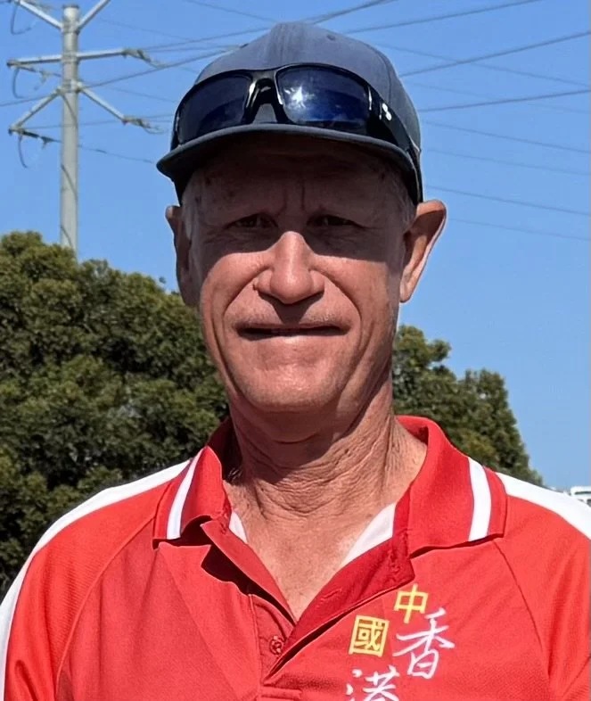 A man wearing a black cap with sunglasses on top, red sports shirt with Chinese characters, standing outdoors with trees and power lines in the background.