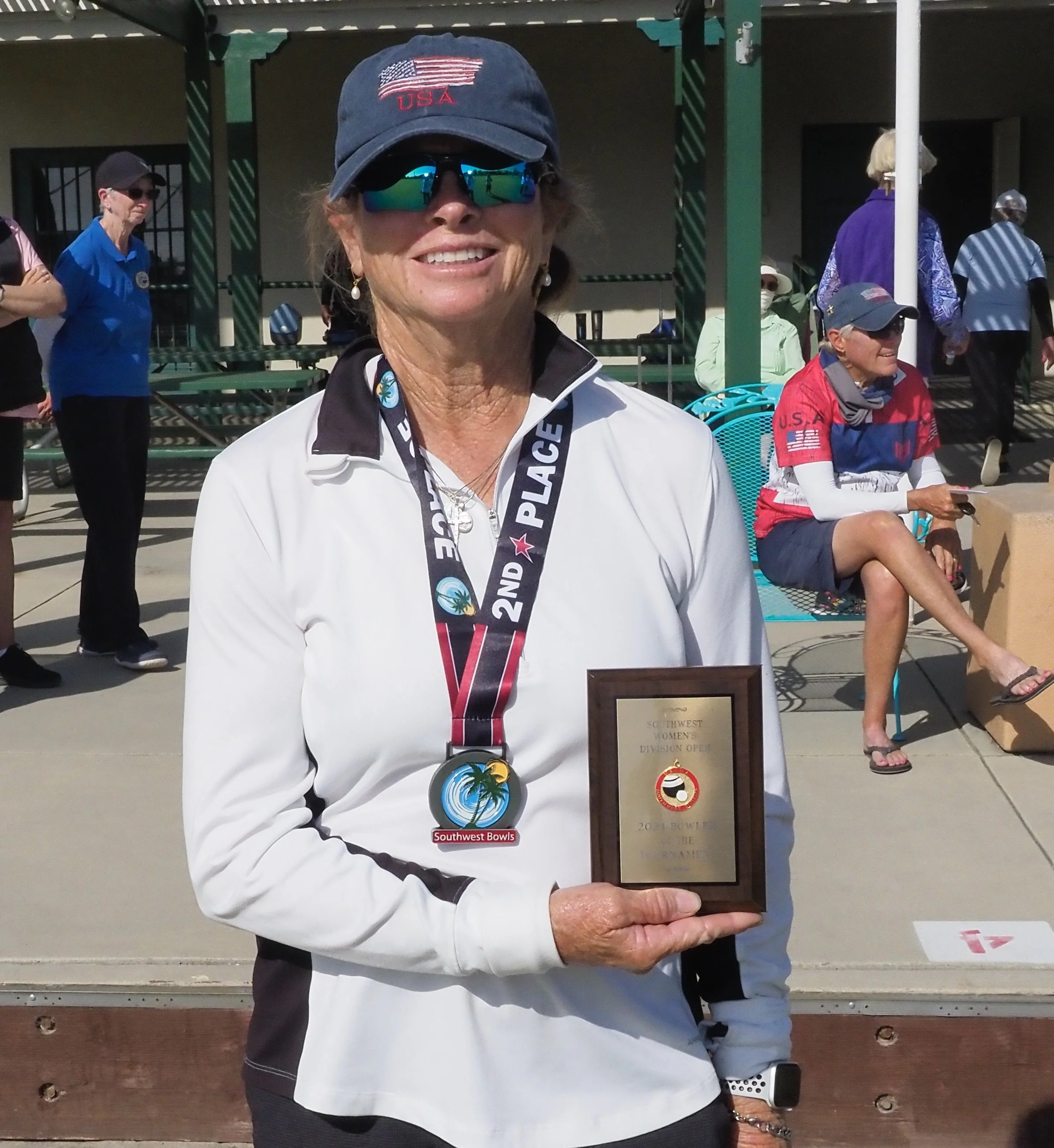A woman wearing a USA cap, sunglasses, and a white and black jacket holding a plaque and medal, smiling at the camera, at an outdoor event with other people in the background.