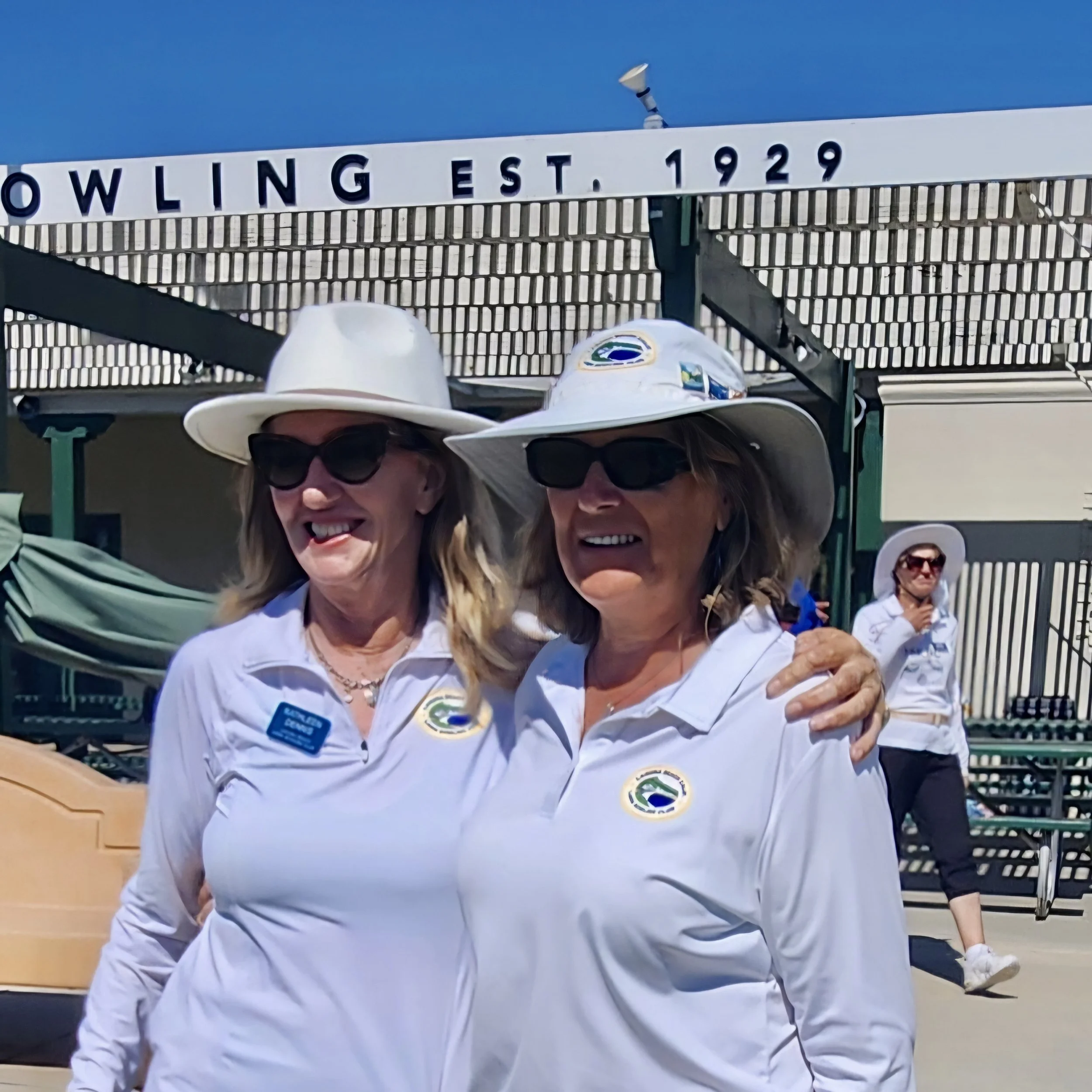 Two women wearing white shirts, hats, and sunglasses standing outdoors in front of a building with a sign that reads "Bowling EST. 1929," smiling and posing for the photo. One woman has her arm around the other.