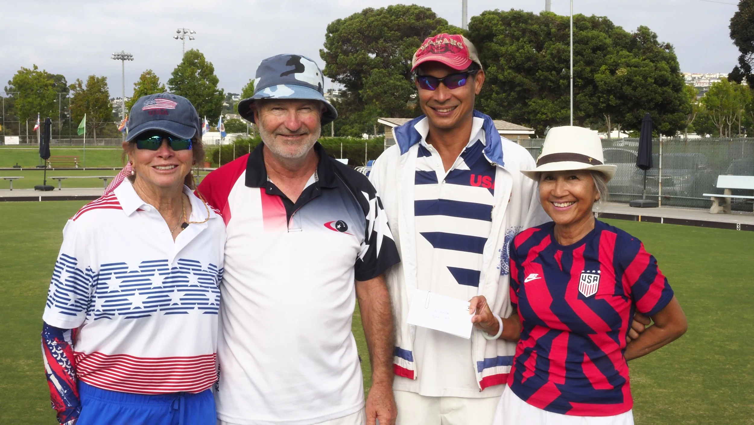 Four people wearing patriotic USA sports attire, standing on a grass sports field, smiling at the camera.