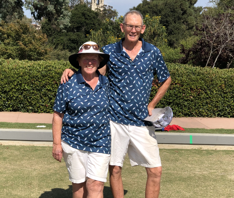 A smiling older woman and man wearing matching blue shirts with white paisley pattern, white shorts, and sunglasses standing on a grass lawn outdoors. They are close together with their arms around each other, with greenery, a pathway, and trees in the background.