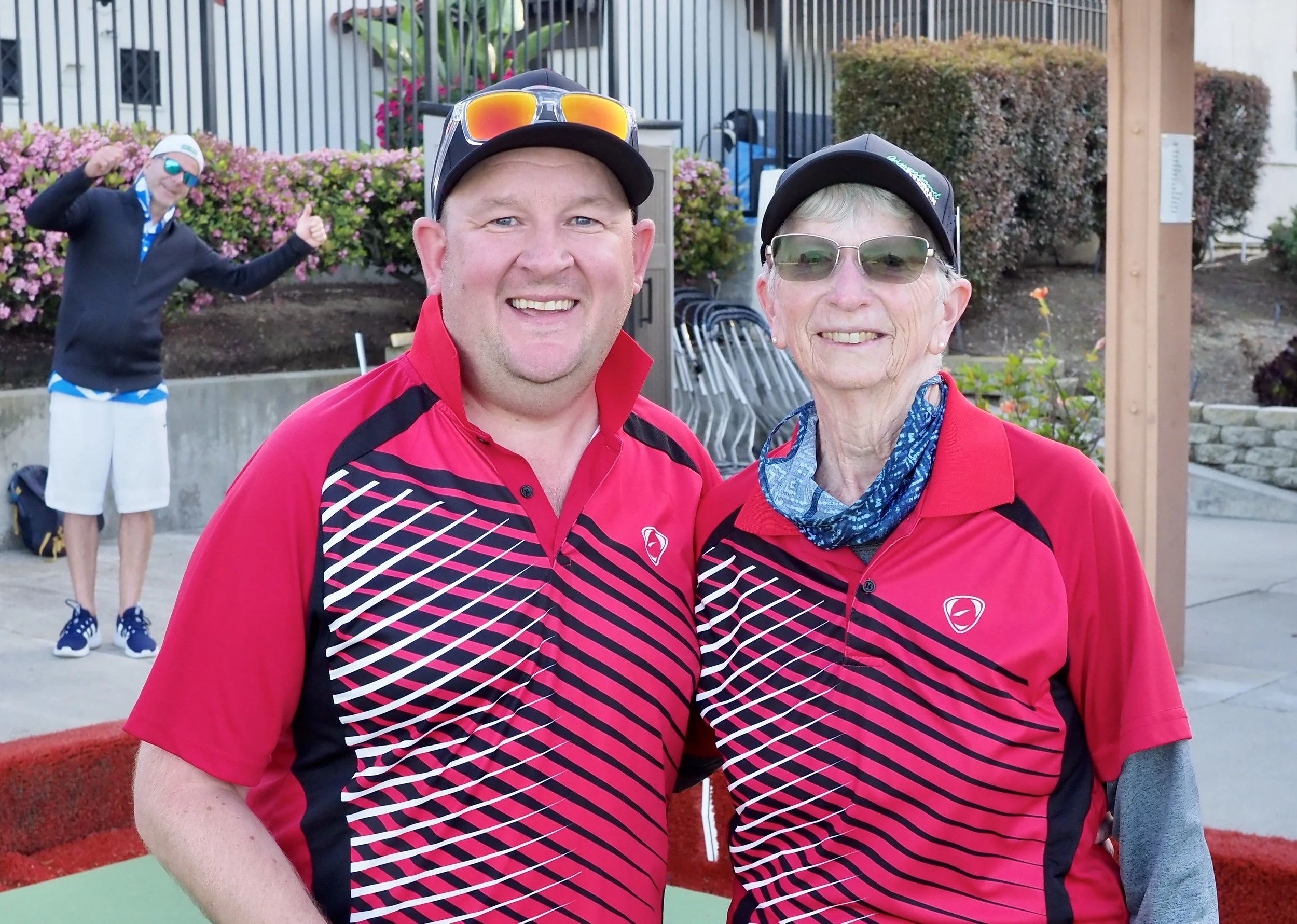 Two men and a woman wearing matching red and black sports jerseys, taking a selfie outdoors with a third person in the background celebrating and giving thumbs-up.