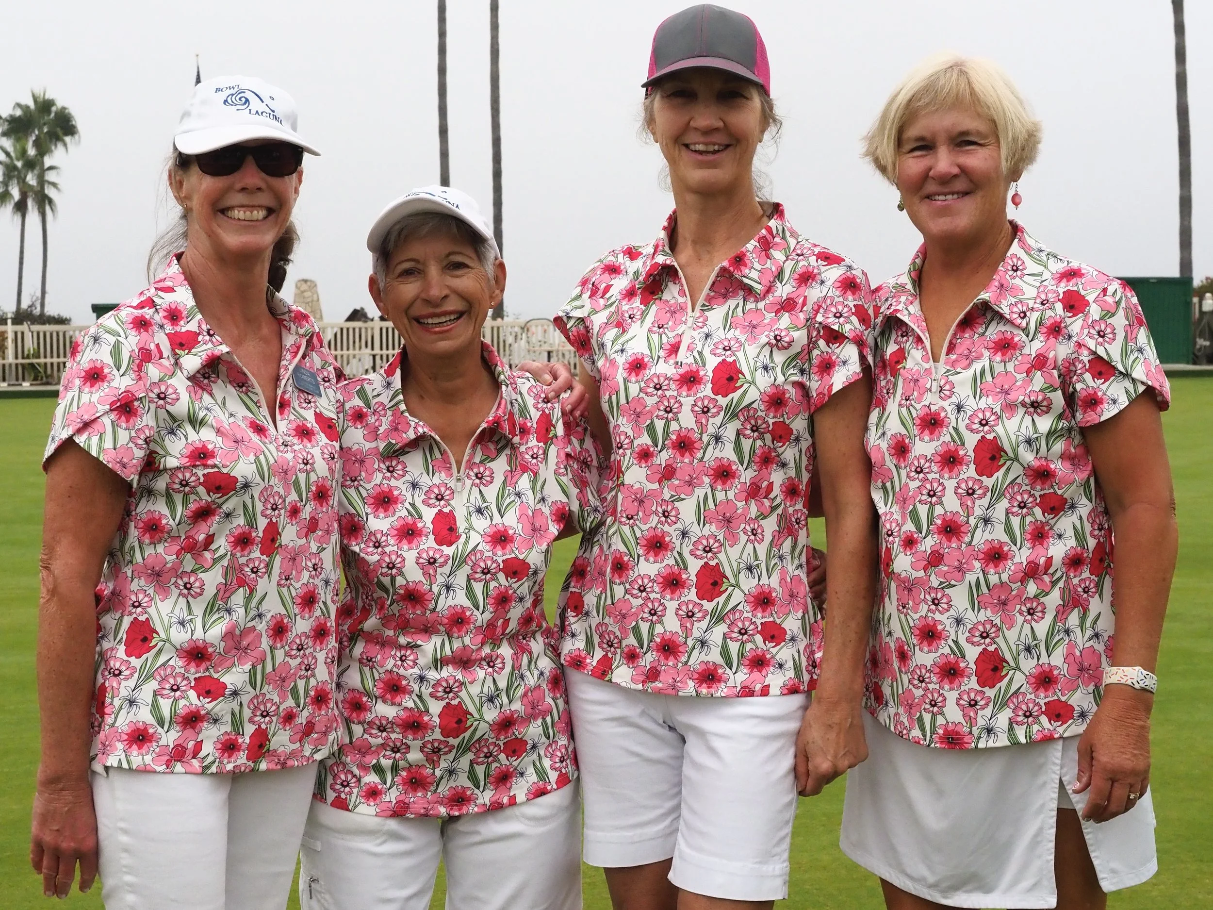 Four women wearing matching pink, red, and white floral shirts and white skirts or shorts, standing on a golf course, smiling for the camera.