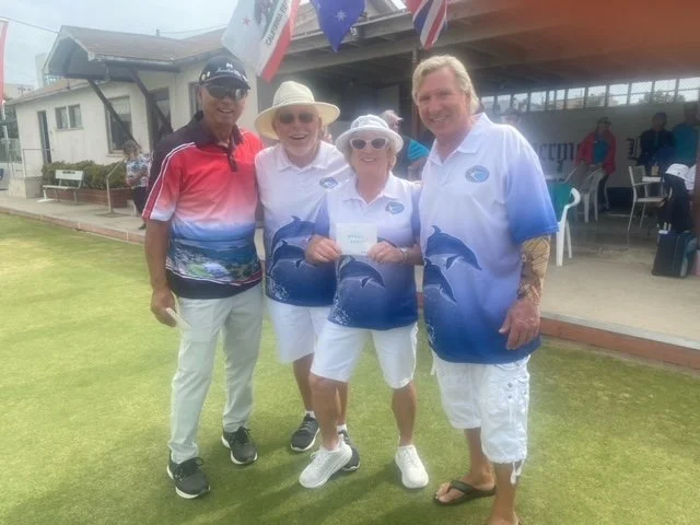 Four people standing on a lawn, smiling, with a building and flags in the background. They are dressed in casual and matching custom shirts, some with dolphin designs.