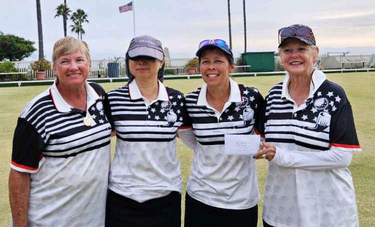 Four women wearing matching black, white, and patriotic-themed sports shirts standing on a green lawn, smiling, with an American flag and palm trees in the background.