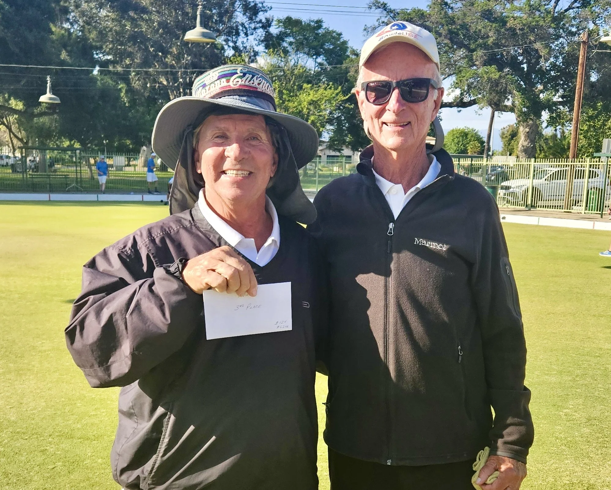 Two older adults standing on a lawn, smiling, with one holding a white note that says "3rd Place" and the other holding a yellow item, possibly a towel, in a sports setting with a fence, trees, and a few people in the background.