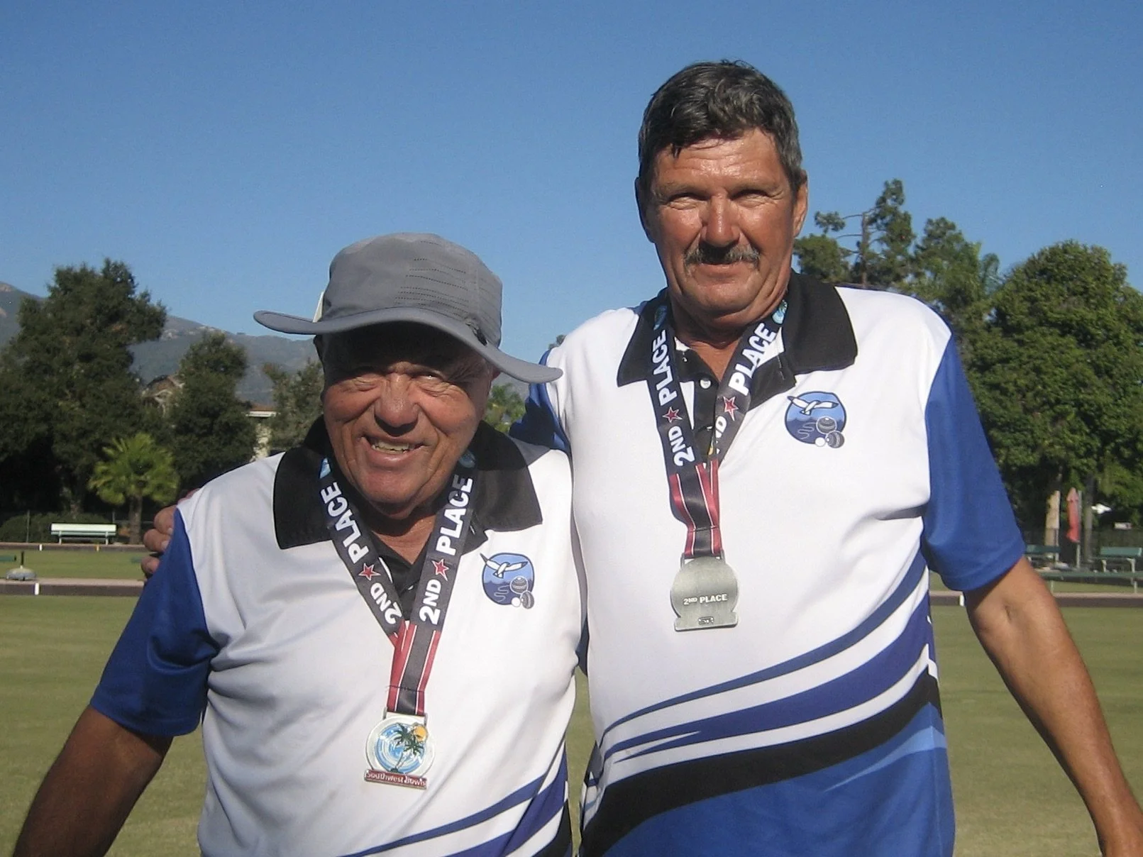Two men wearing white and blue sports polo shirts with medals around their necks, standing on a lawn with trees and mountains in the background, smiling after a sports event.