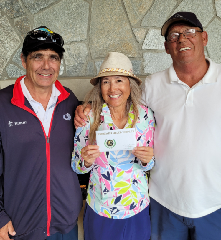 Three smiling people standing together, with the woman in the middle holding a certificate that reads 'Coronado Mixed Triples', against a stone wall background.
