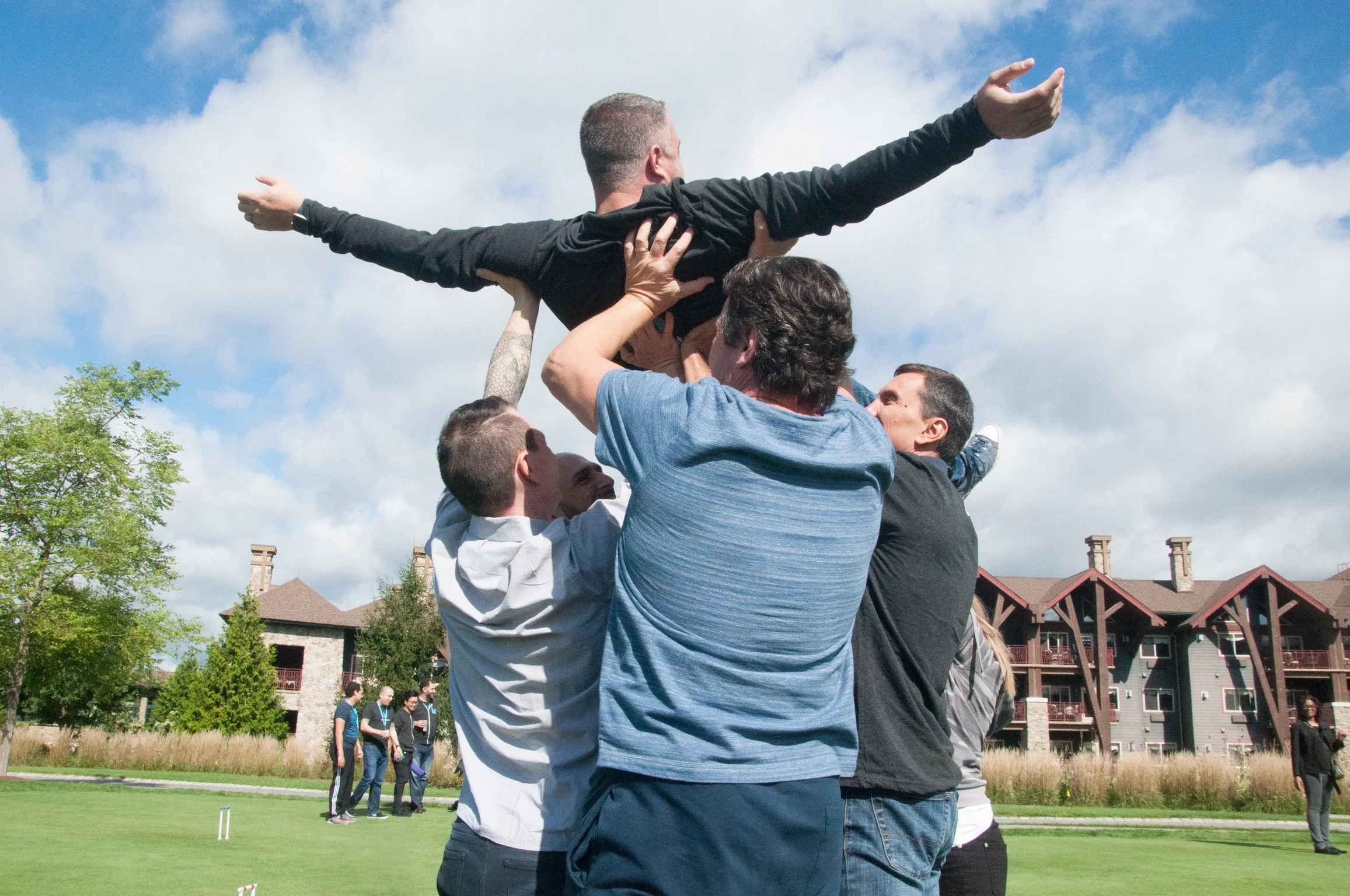 Group of people outdoors celebrating as they lift a person horizontally in the air during a team-building activity on a sunny day.