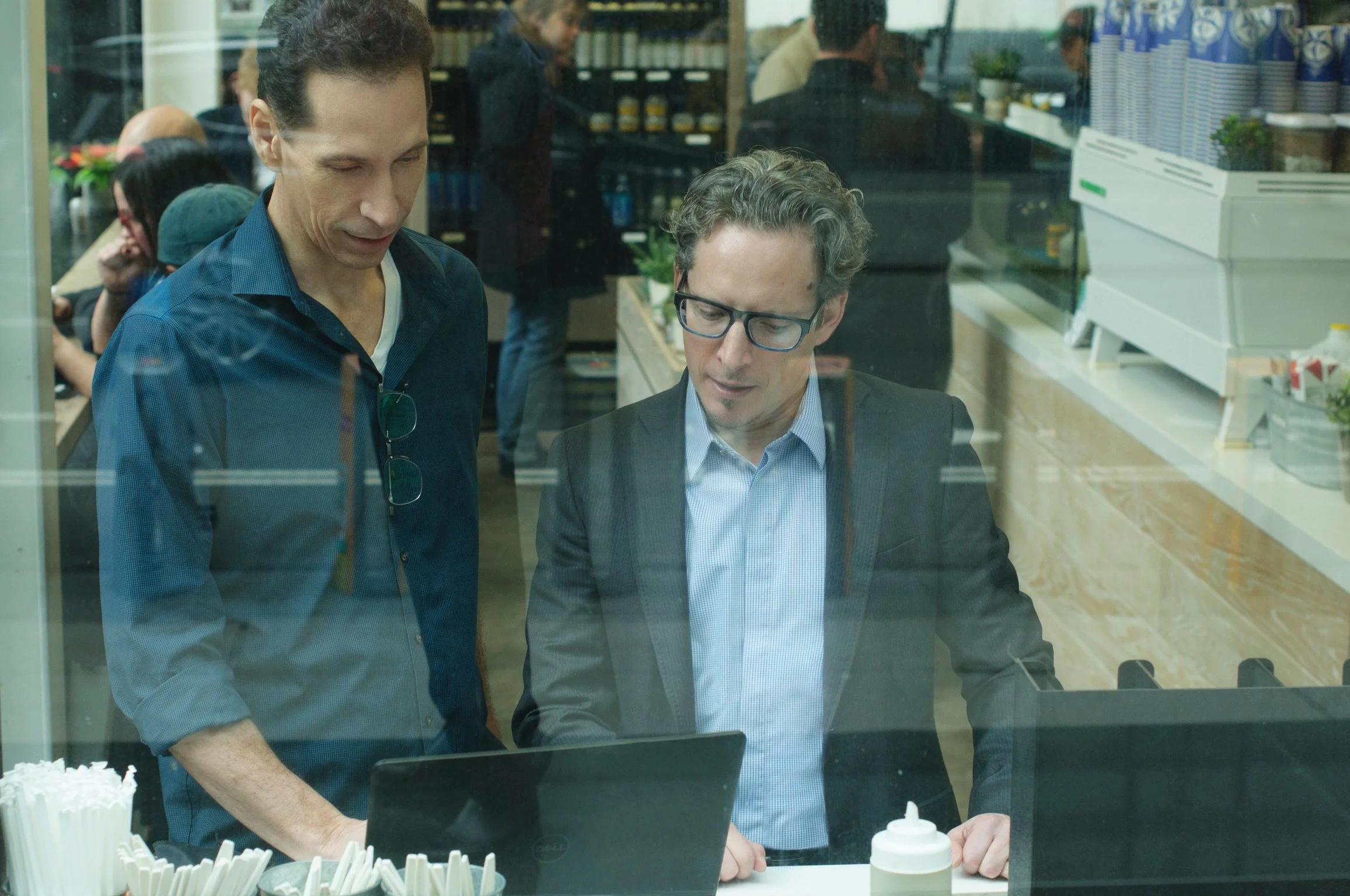 Two men examining a laptop inside a cafe, viewed through a glass window.