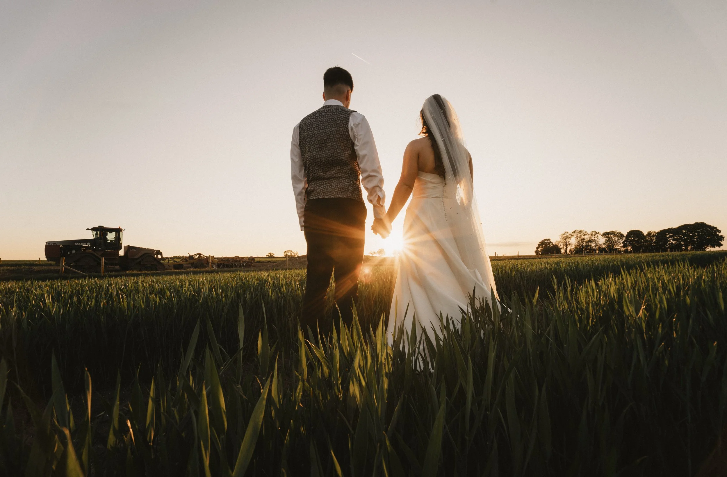 A bride and groom holding hands during sunset in a green field, with farm equipment in the background.