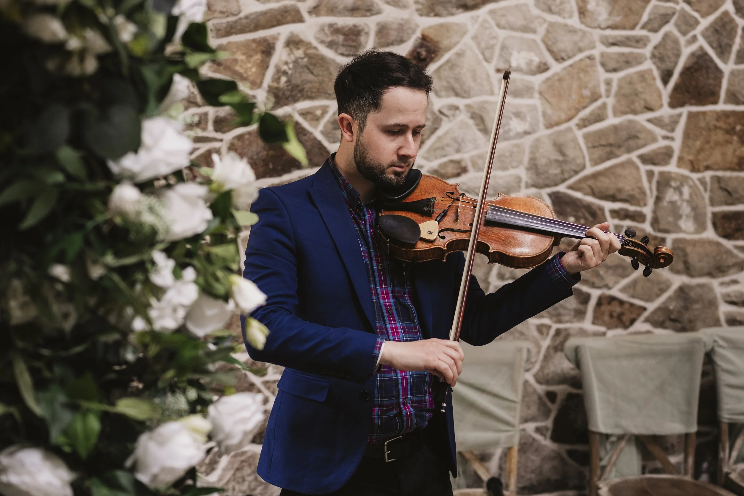 A man in a blue blazer playing the violin in a rustic room with a stone wall background.