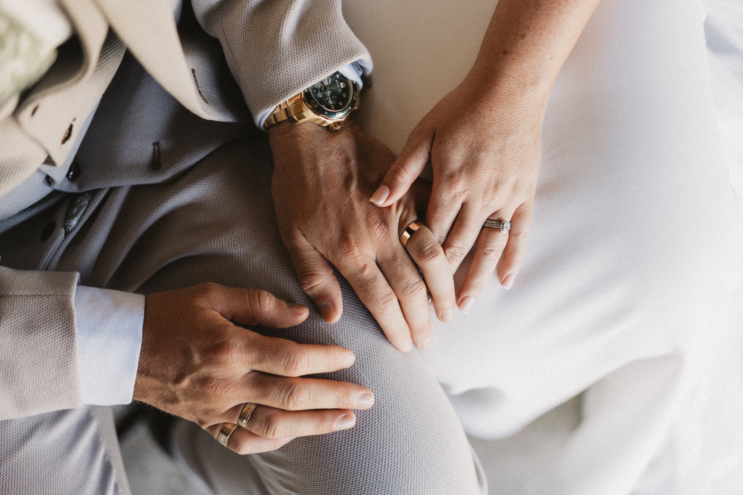 Close-up of a couple's hands layered on top of each other, showing wedding rings. The woman has her hand atop the man's, both are wearing rings, and the woman is wearing a diamond ring and a silver ring, while the man has a gold wedding band. The wom