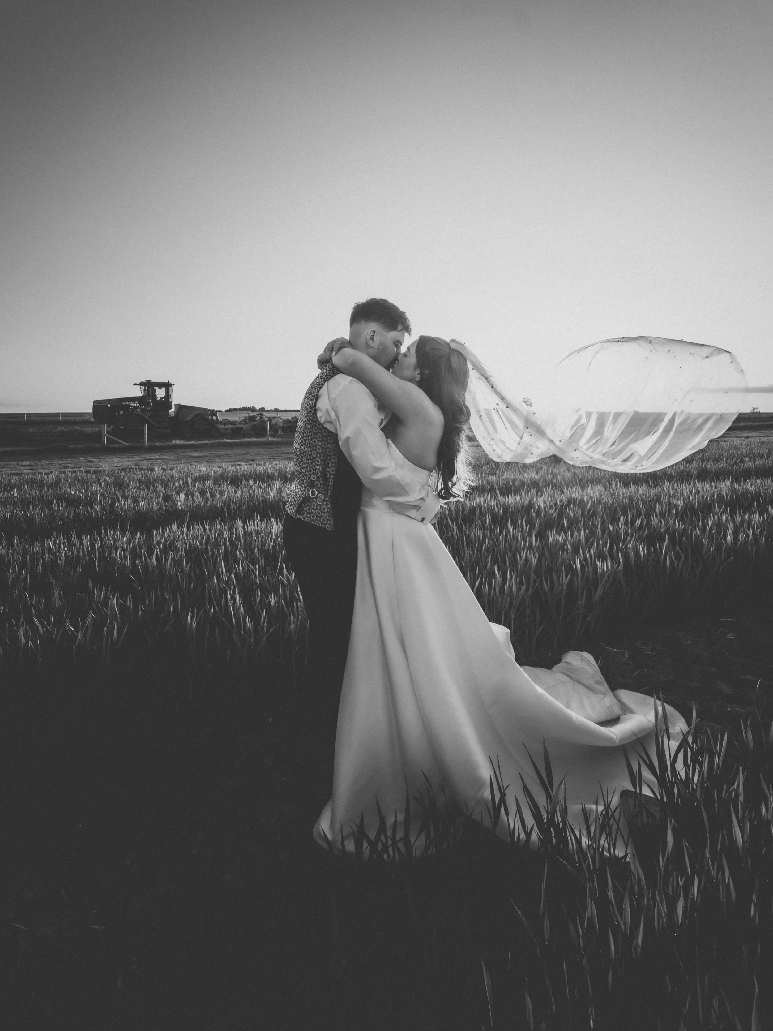 A bride and groom sharing a kiss in a wheat field, with a tractor in the background, during sunset or sunrise.