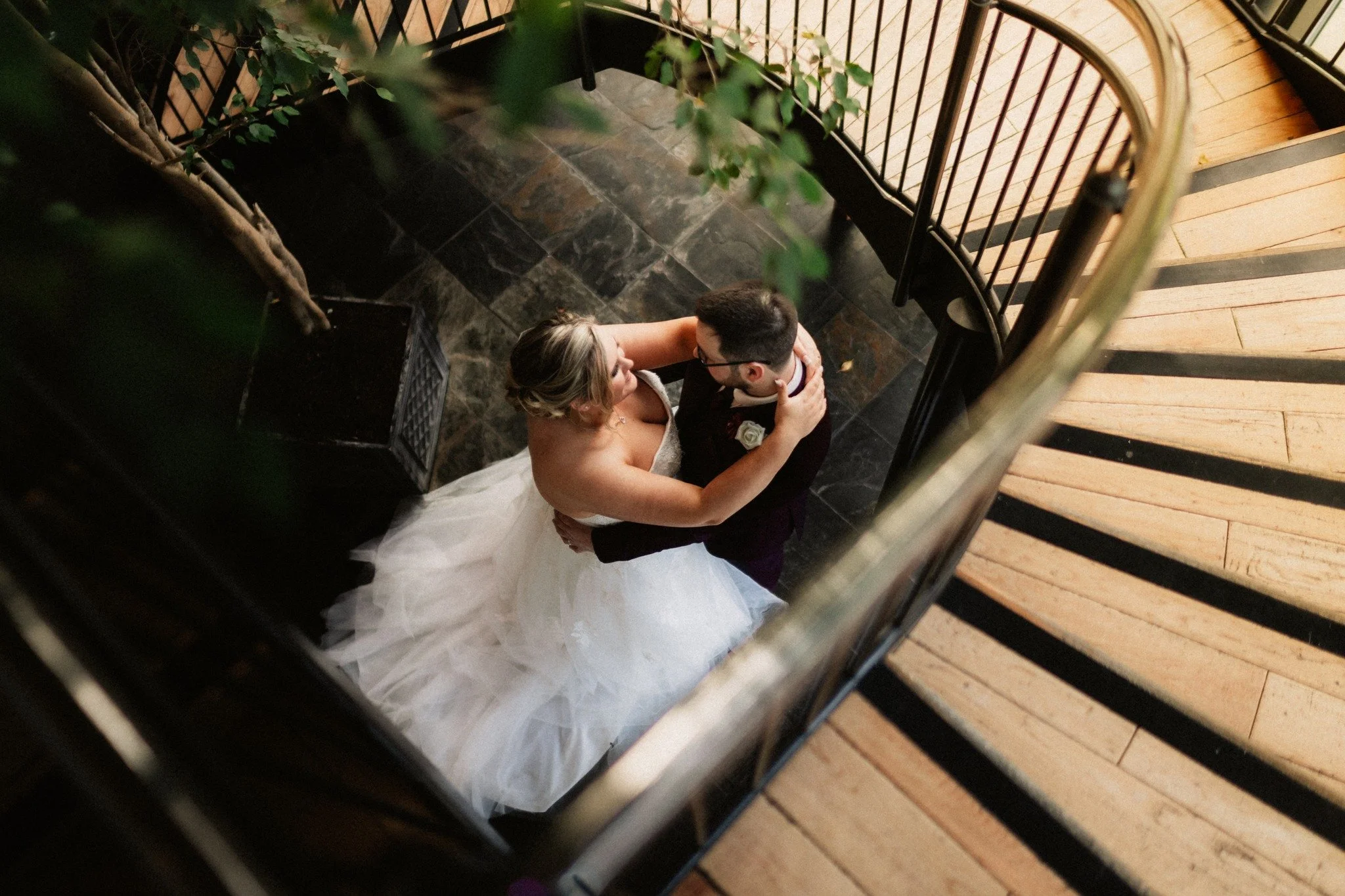 A bride and groom dancing on a spiral staircase, viewed from above, with the bride in a white gown and the groom in a dark suit.