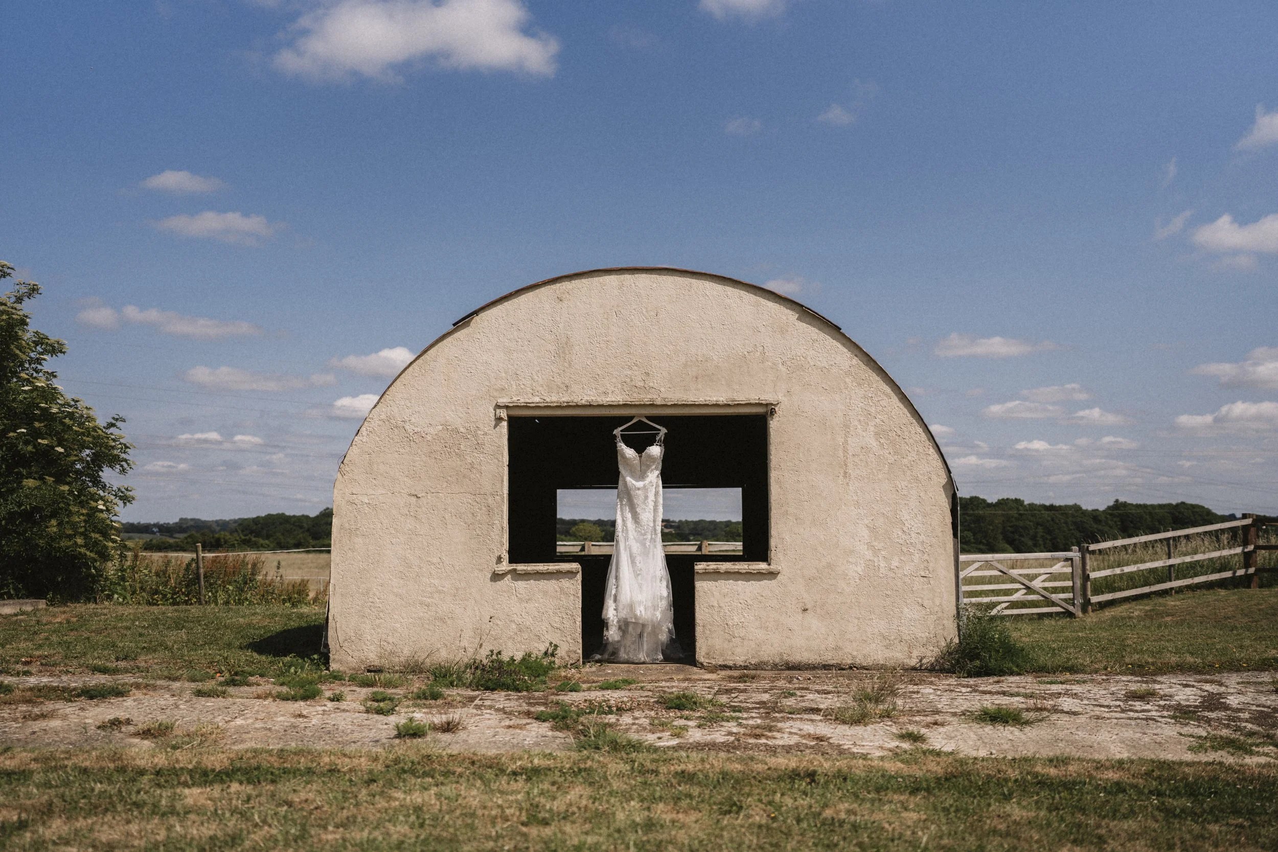 A white wedding dress hanging in a barn window, with a rural field and blue sky in the background.