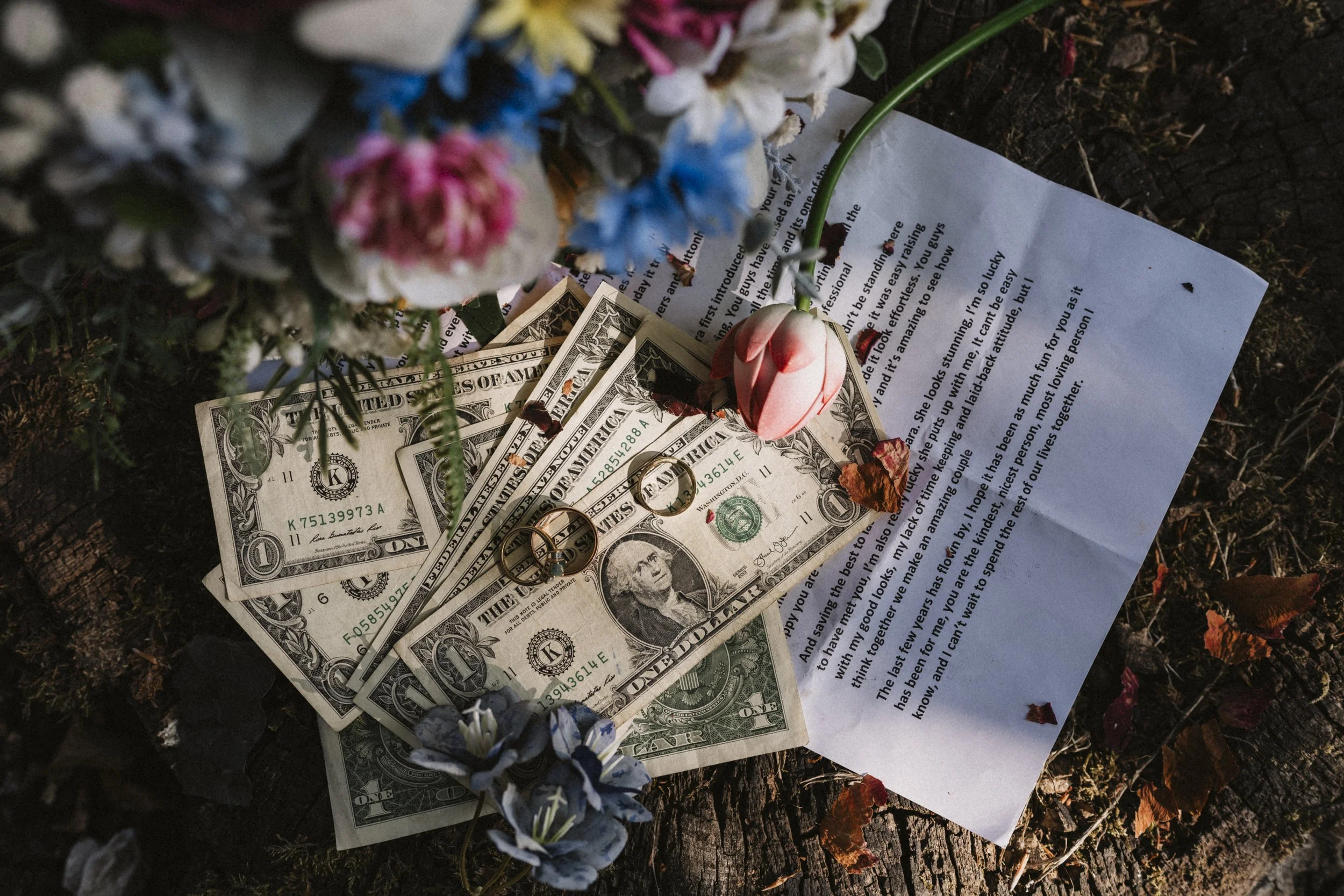 Three one-dollar bills, two gold rings, a pair of earrings, a pink rose, and a letter on a wooden surface with flowers.