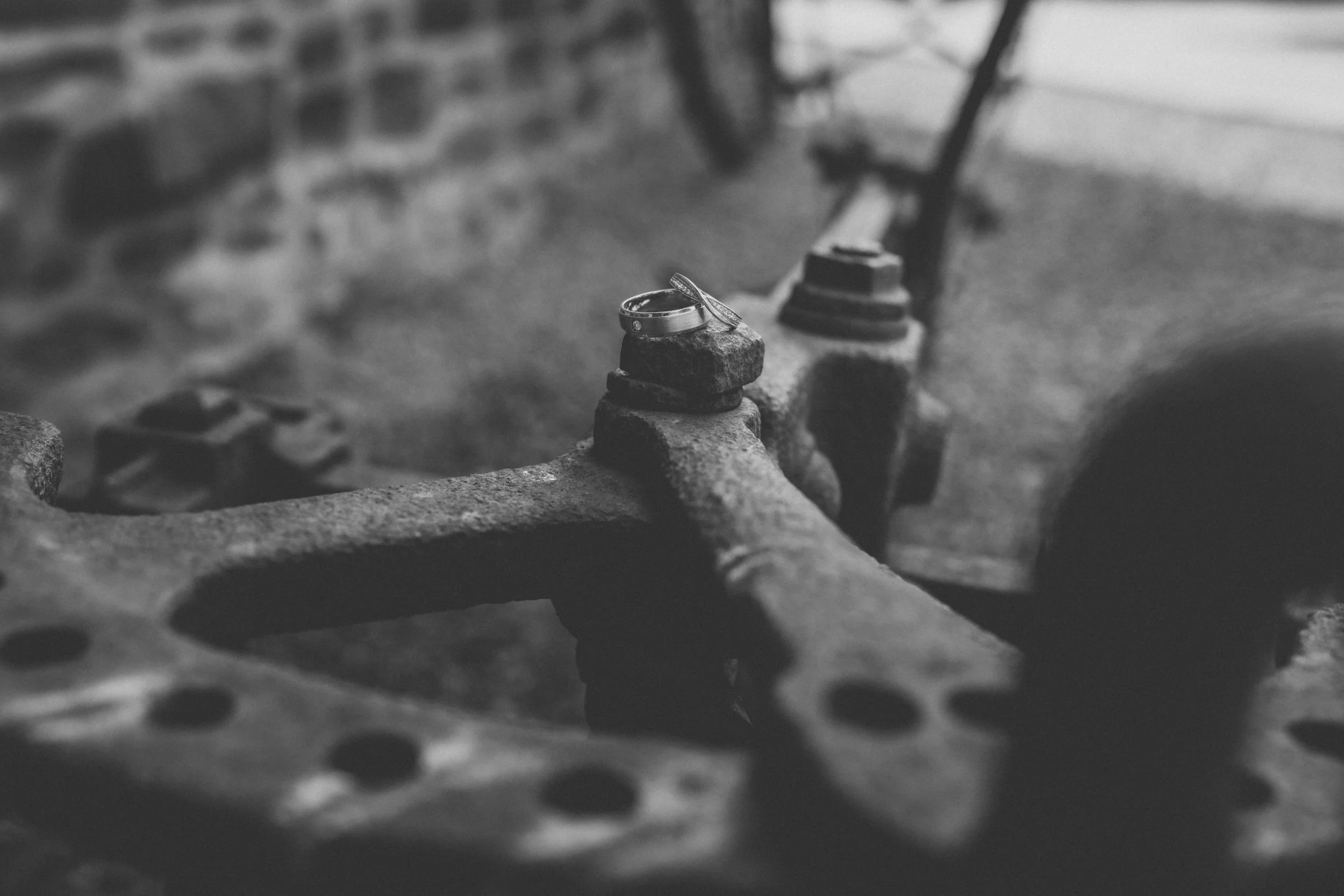 Close-up black and white photo of rings placed on rocks atop a rusty metal surface, with a blurred background of a brick wall and a bench.