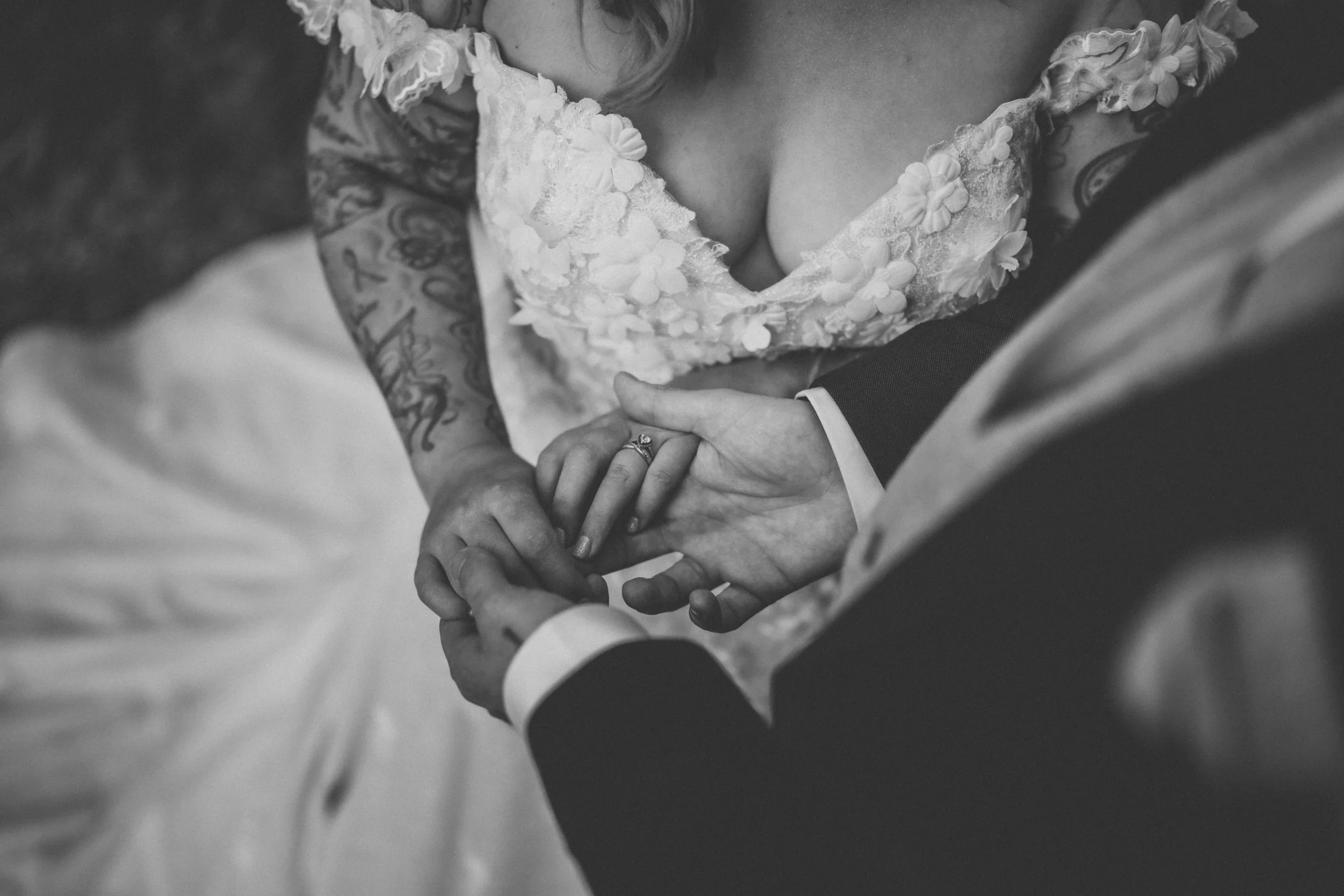 Black and white photo of a bride and groom holding hands during their wedding ceremony, focusing on their intertwined fingers and the bride's wedding ring.