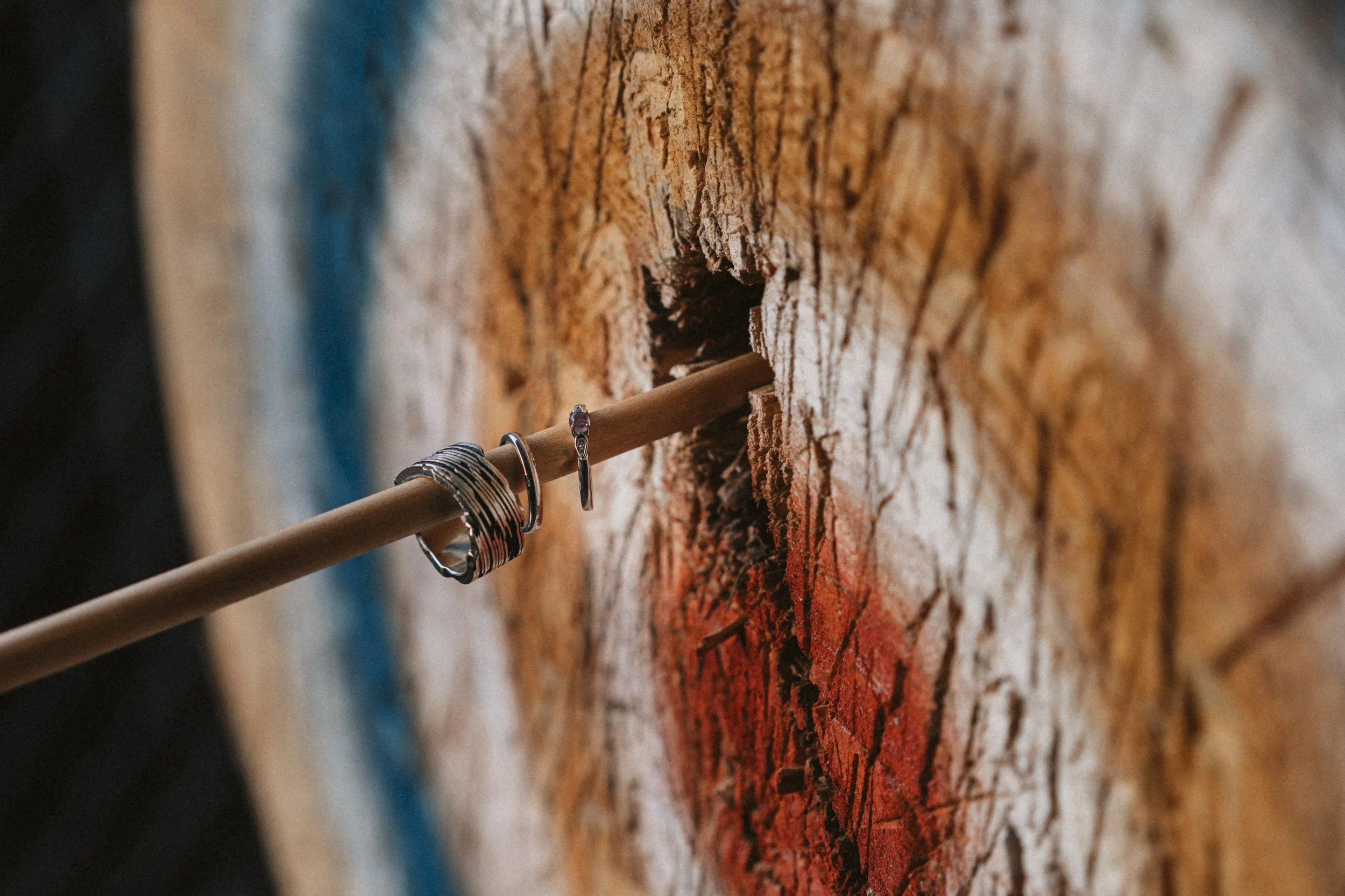 Close-up of a dartboard with a wooden dart stuck in the bullseye, which is marked with red and black rings, and jewelry including rings and bracelets hanging from the dart.