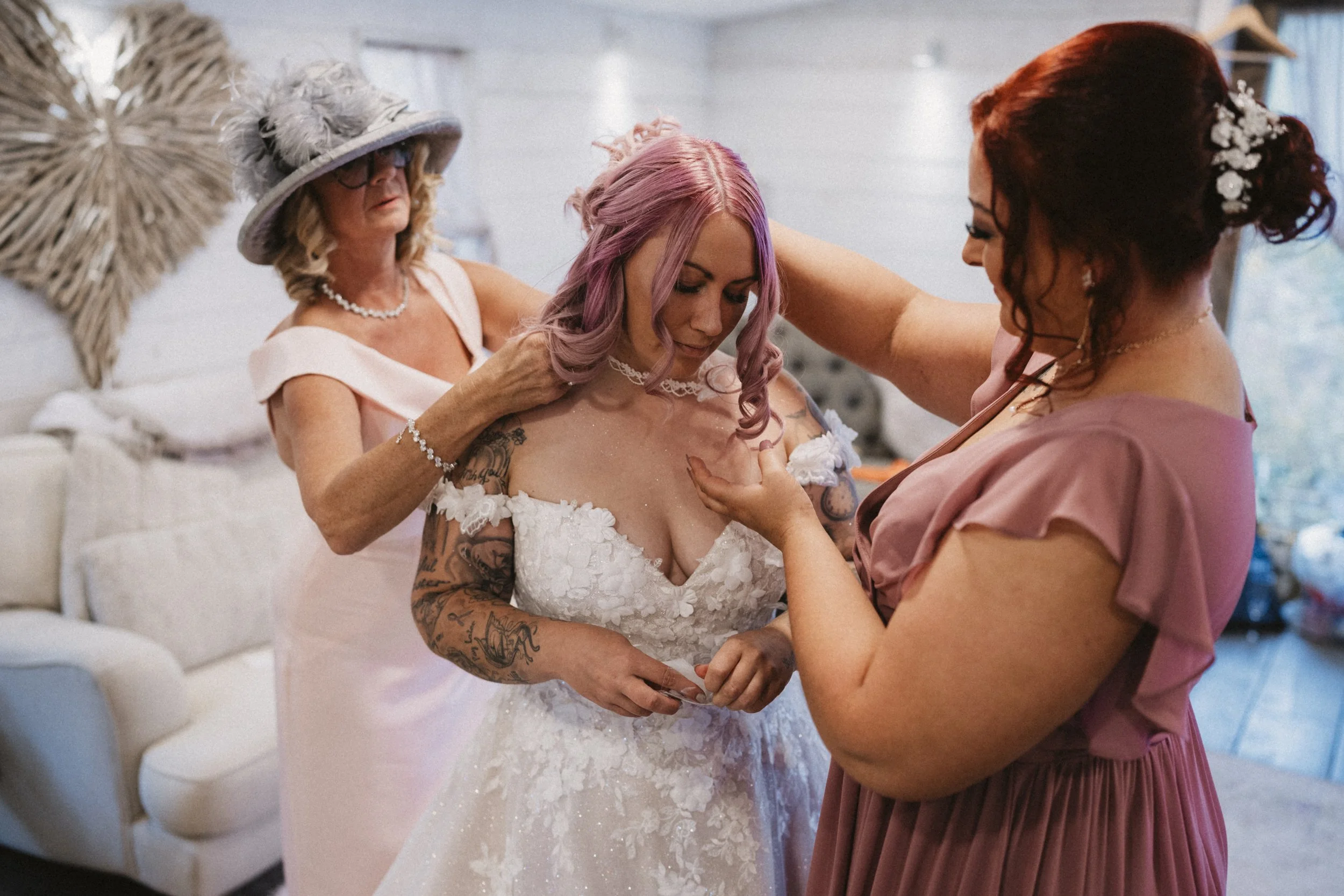 A bride with pink hair in a white wedding dress is being helped by two women, one with red hair and the other with blonde hair and a hat, in a well-lit room decorated with a large wicker heart on the wall.