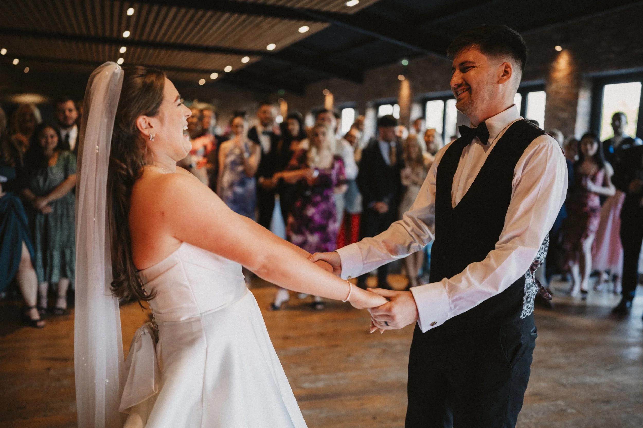 A bride and groom dancing at their wedding reception, holding hands and smiling at each other, with wedding guests dancing in the background inside a decorated venue.