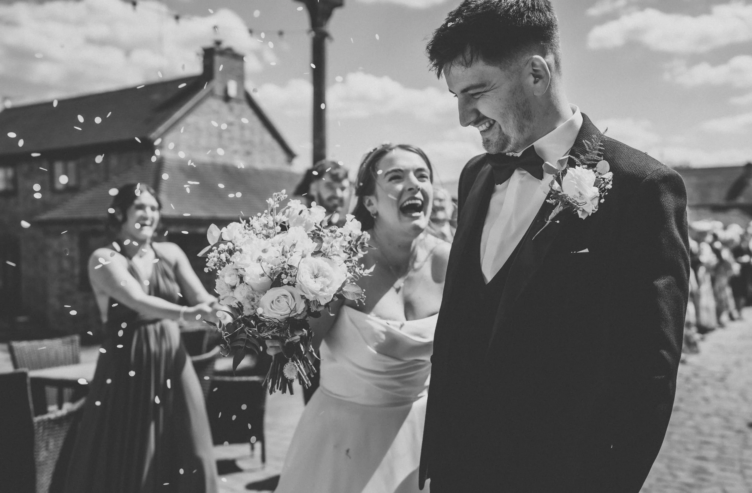 A wedding celebration with a bride and groom smiling and celebrating outdoors, with friends throwing confetti.