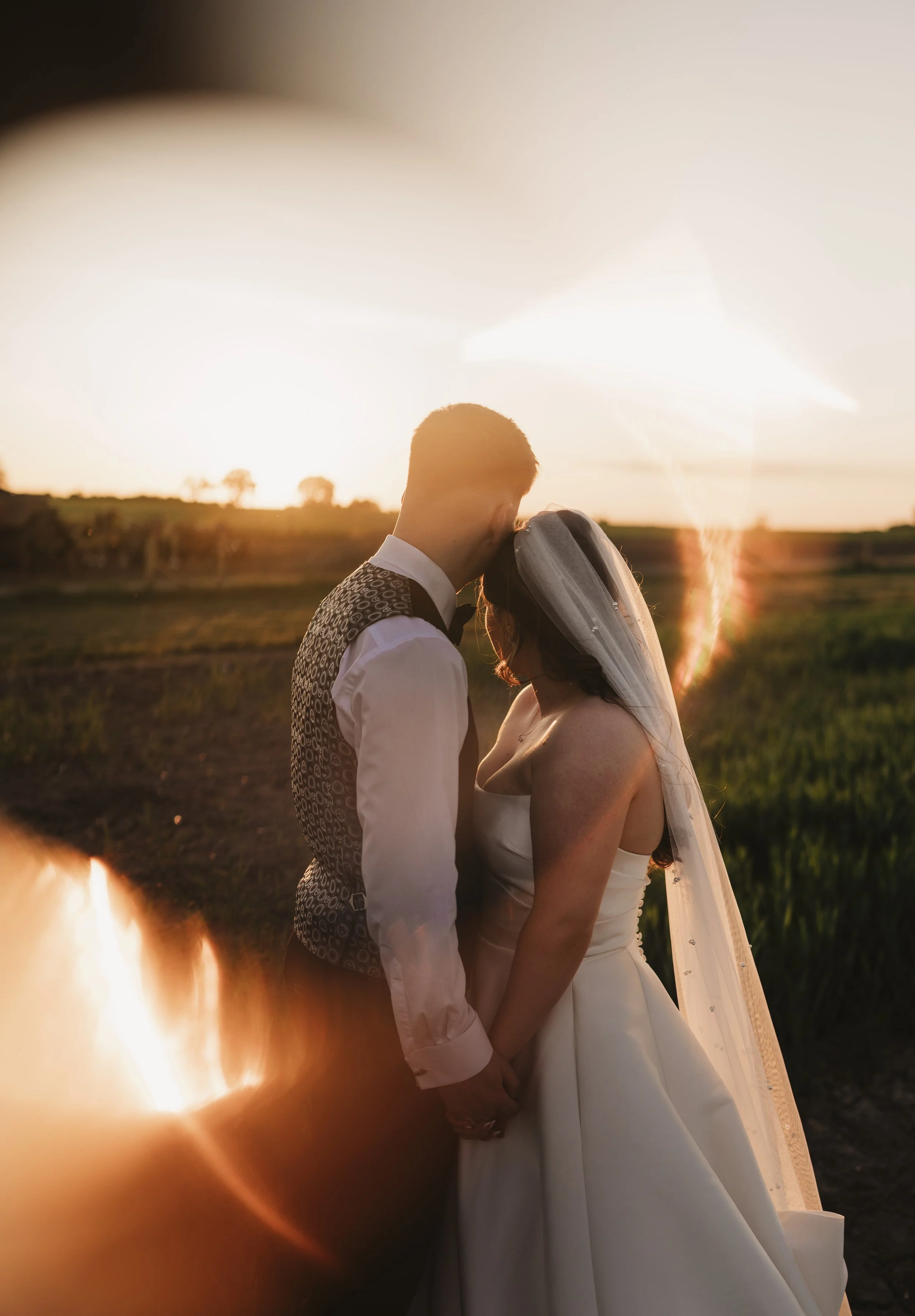 A bride and groom stand close together, holding hands, with their foreheads touching during sunset in a rural field.