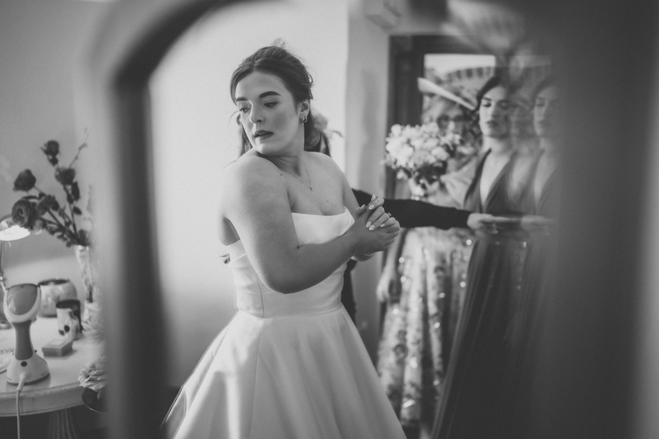 A woman in a wedding dress looking at herself in a mirror, with flowers and other women in the background.