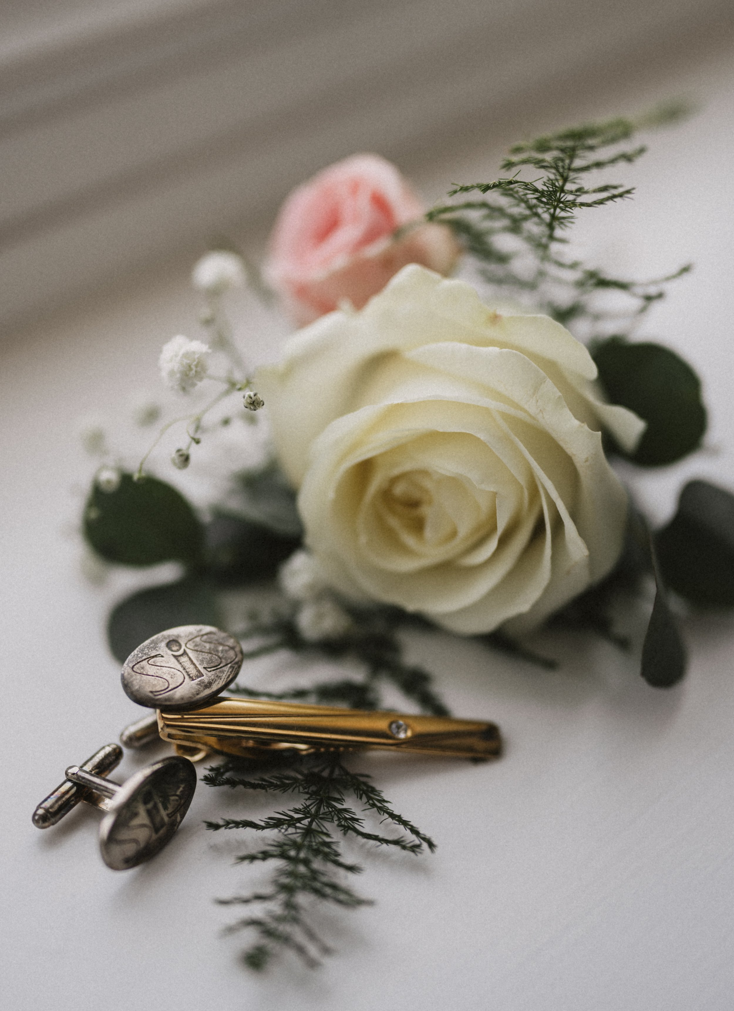 A white rose with greenery and small flowers, a pink rose in the background, and a pair of silver cufflinks with the initials 'SIS' engraved on them.