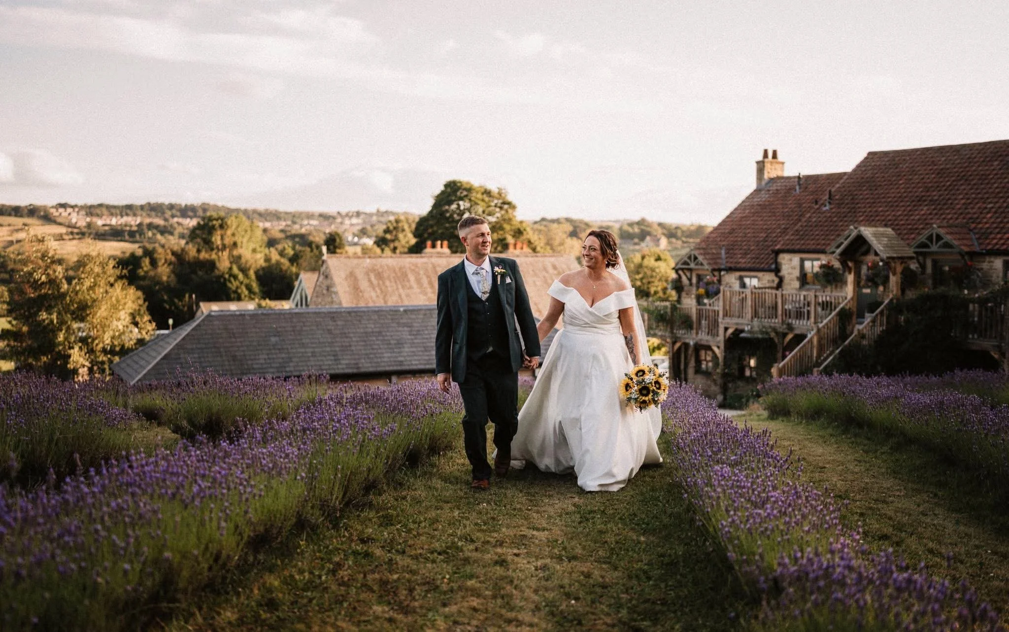 A bride and groom walking hand-in-hand through a lavender field during their wedding. The bride is holding a bouquet of sunflowers and wearing a white off-the-shoulder wedding dress. The groom is in a dark suit and tie. They are smiling at each other