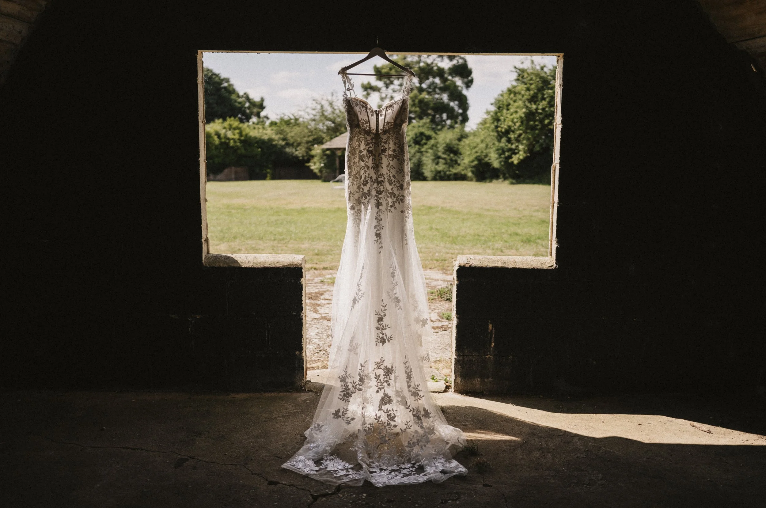 A wedding dress hanging on a hanger outside a dark building, with a grassy field and trees in the background.