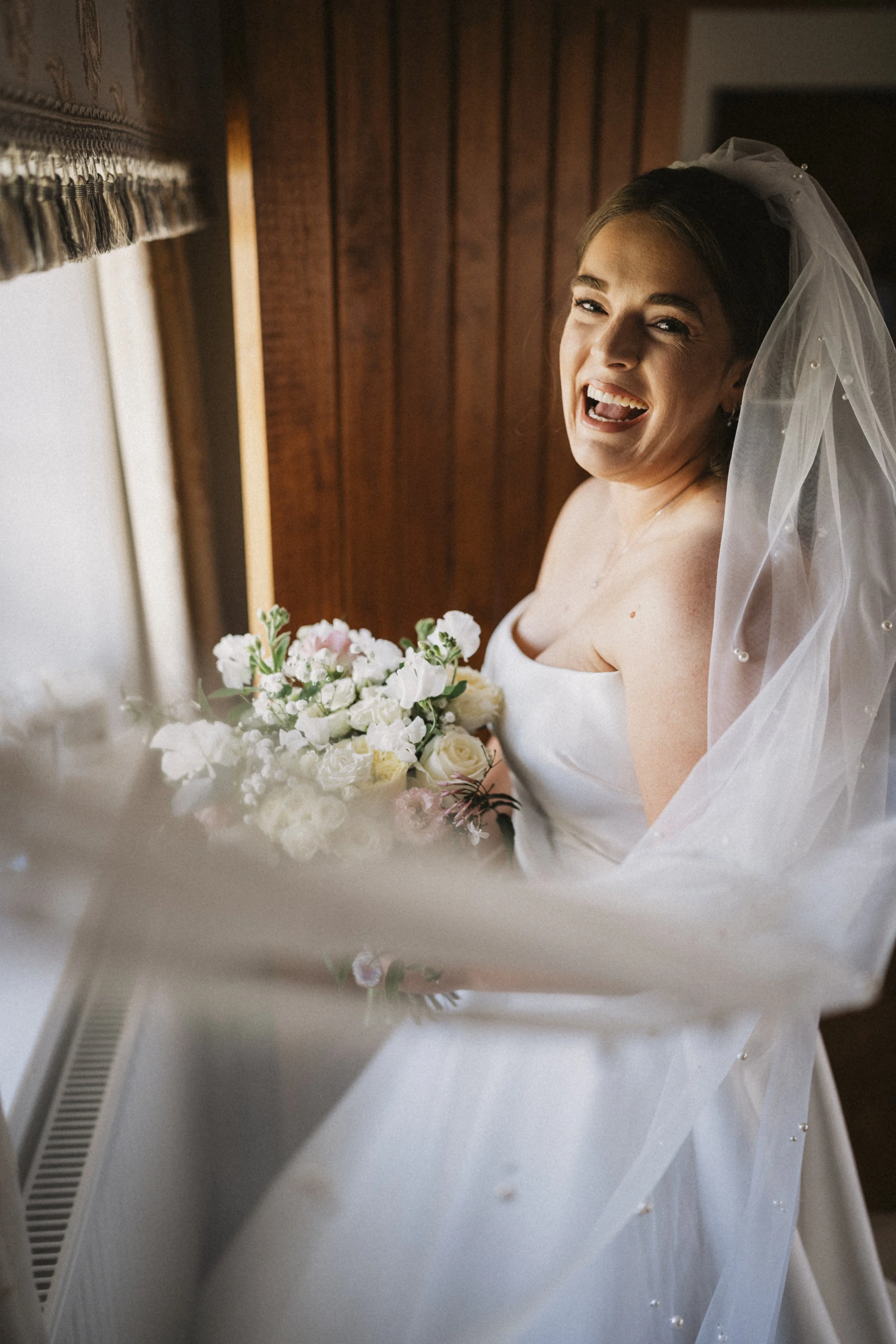A smiling bride in a wedding dress and veil, holding a bouquet of white and pink flowers, standing indoors near a window with wooden paneling behind her.