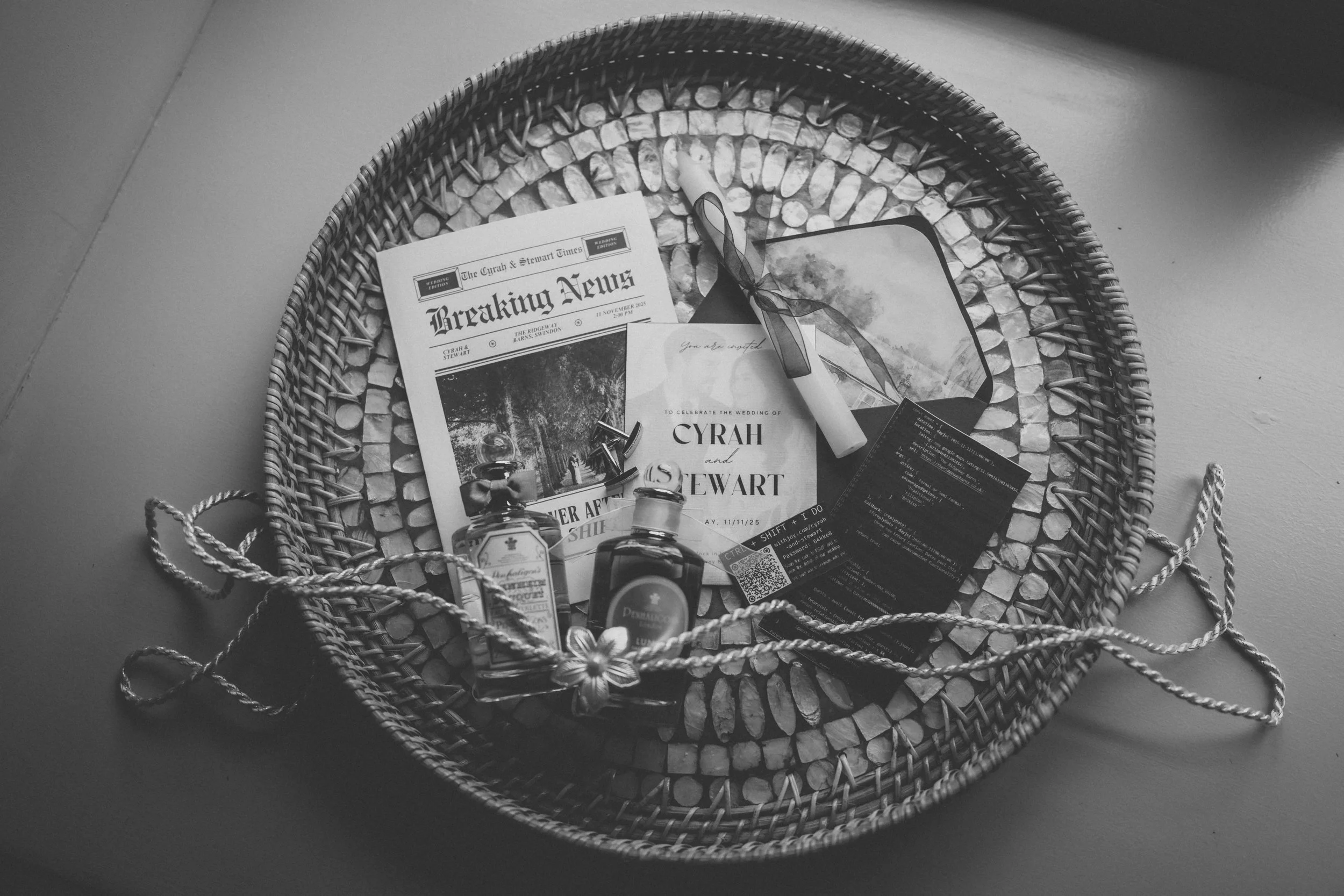A round woven tray with wedding items including a newspaper, wedding invitation, a small jar with ribbon, perfume, a car key, a QR code, and some cards.