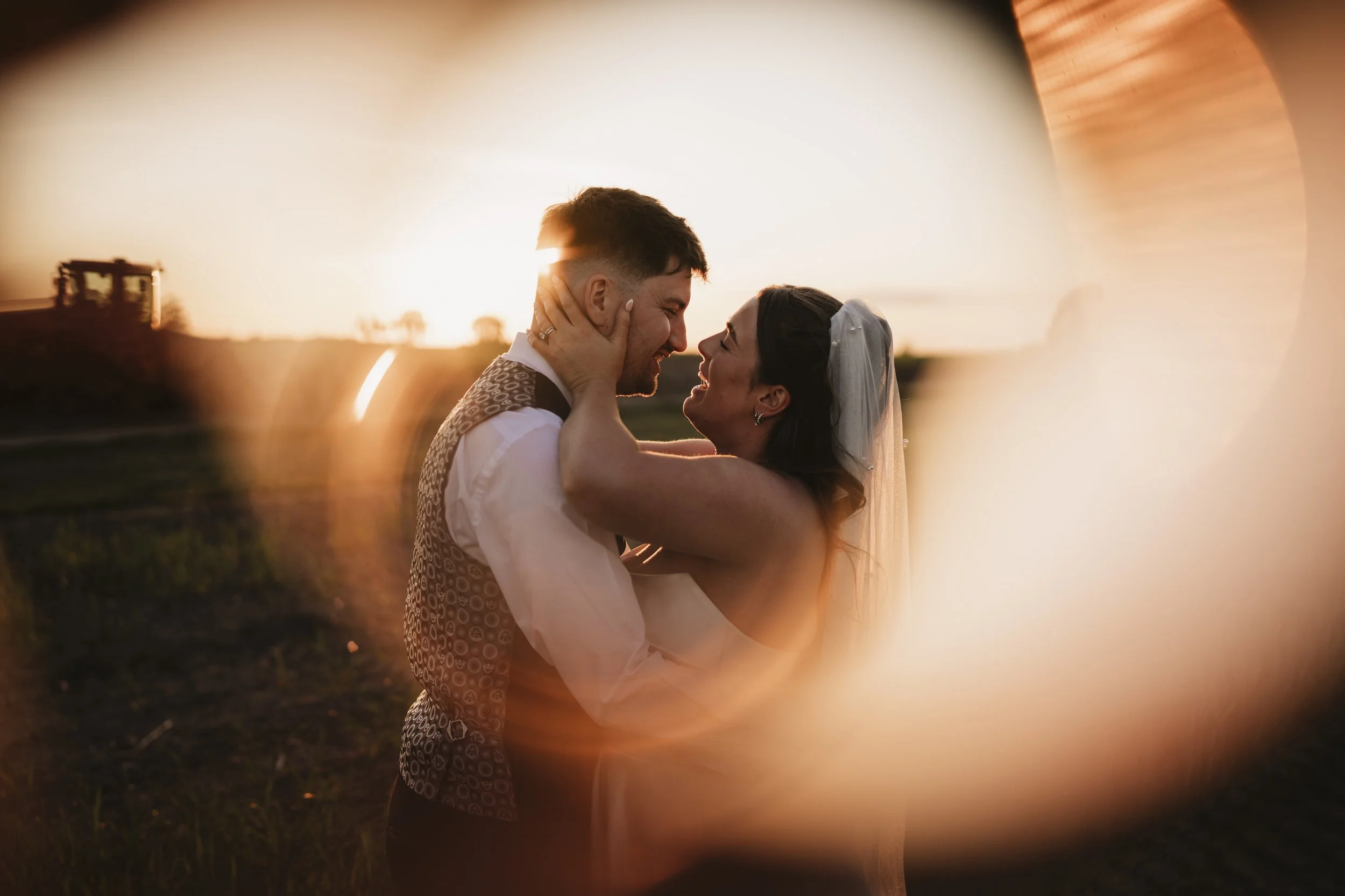 A couple in wedding attire embracing during sunset with the sun setting in the background.