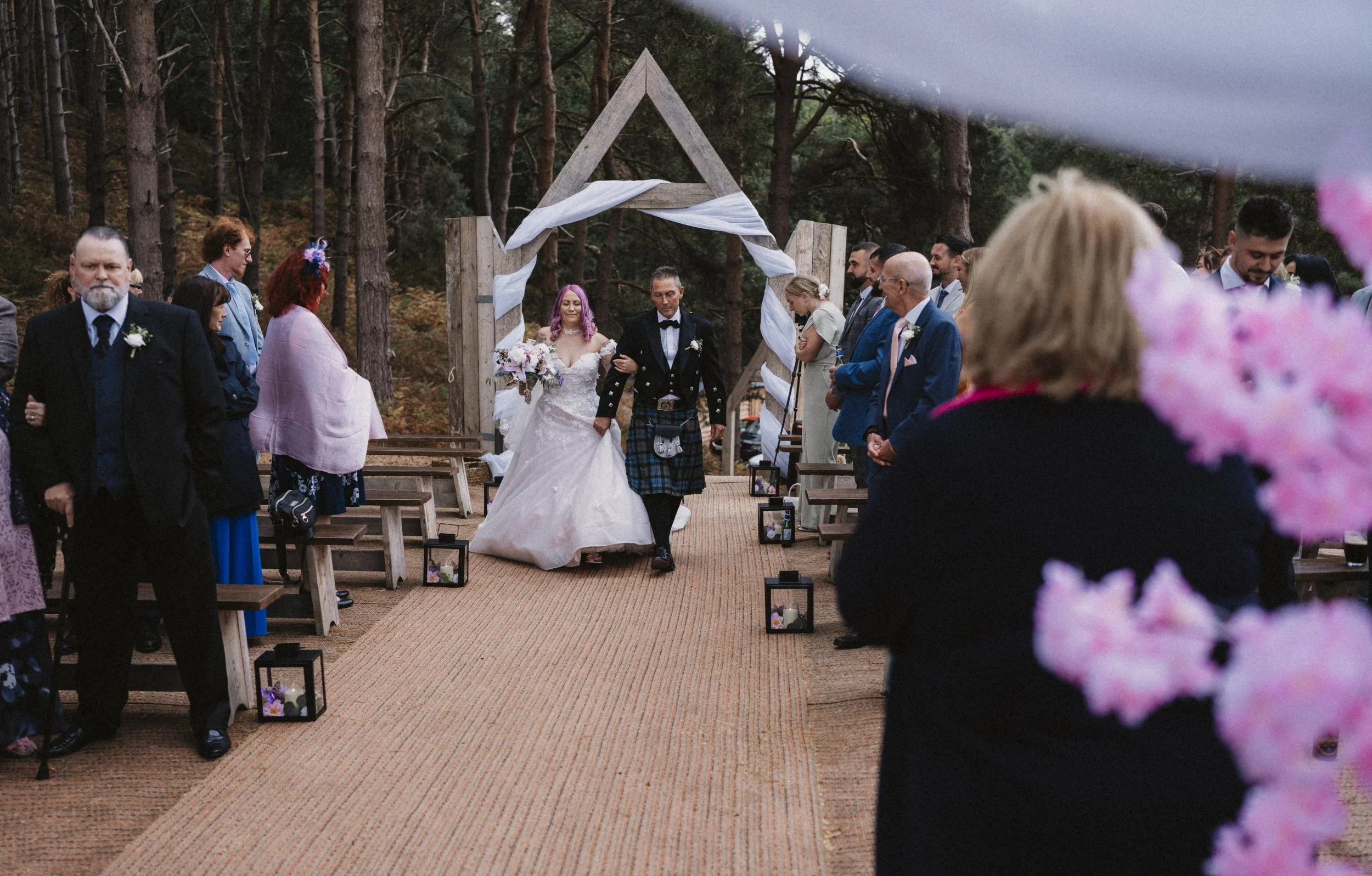 Bride and groom walking down the aisle at an outdoor wedding in a forest, surrounded by guests.