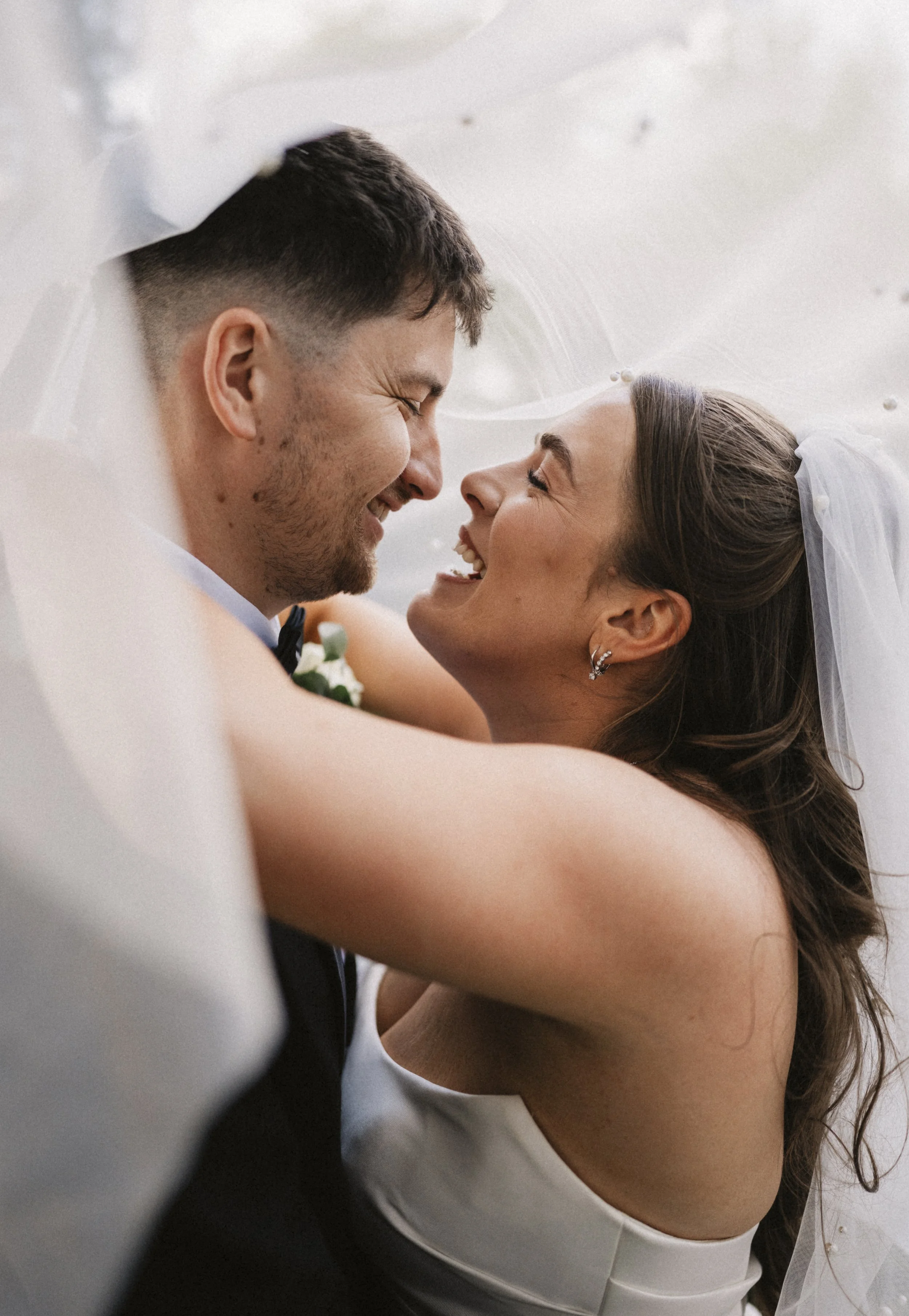 A joyful bride and groom embrace under a veil, smiling with their foreheads touching.