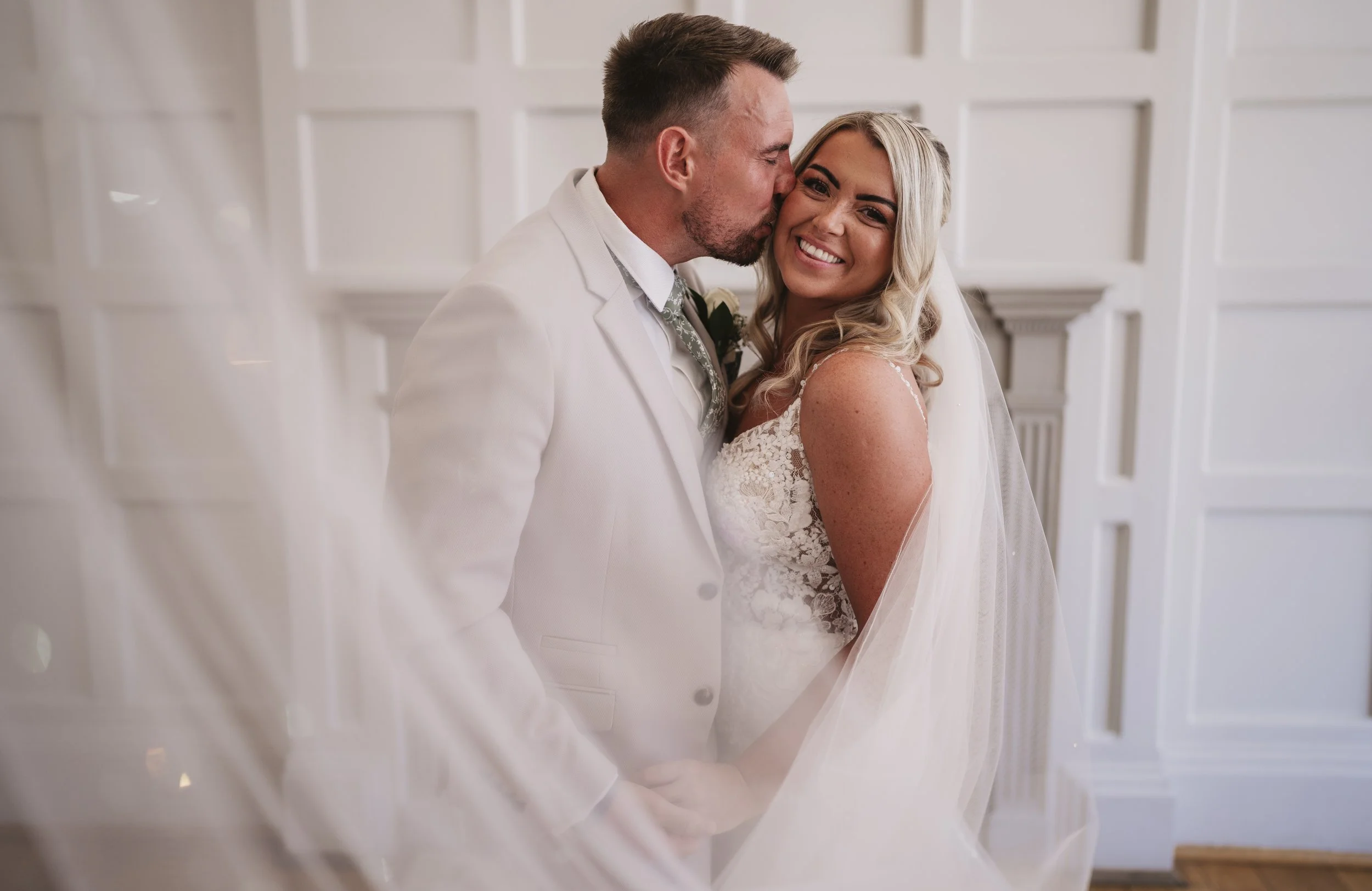 A groom in a white suit kisses a smiling bride in a white lace wedding dress on the cheek in a bright, elegant room.