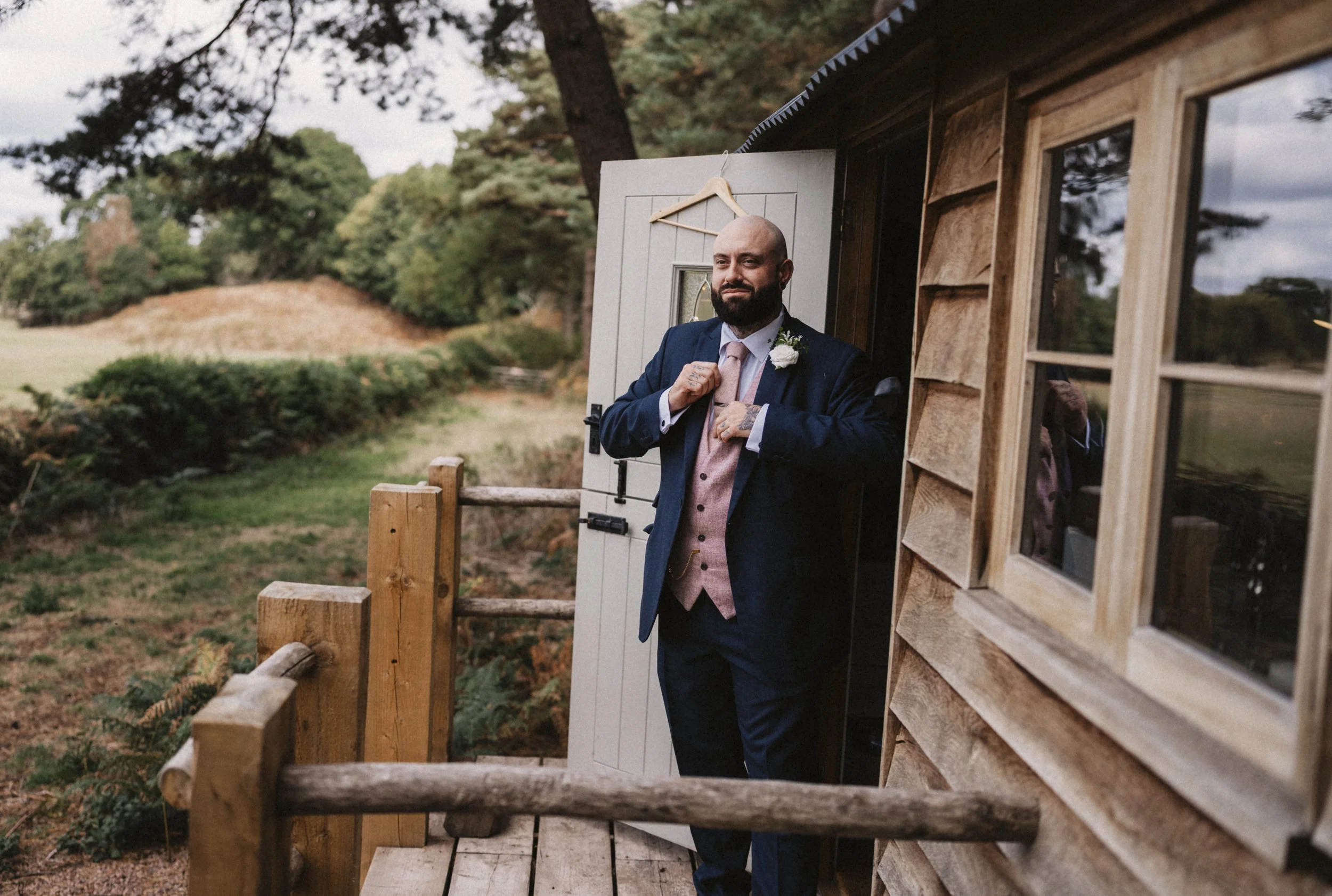 A man dressed in a navy blue suit with a light pink vest and tie, standing on a wooden porch of a rustic cabin with a window, adjusting his suit jacket, with a white flower boutonniere on his lapel, in an outdoor setting with trees and shrubbery.