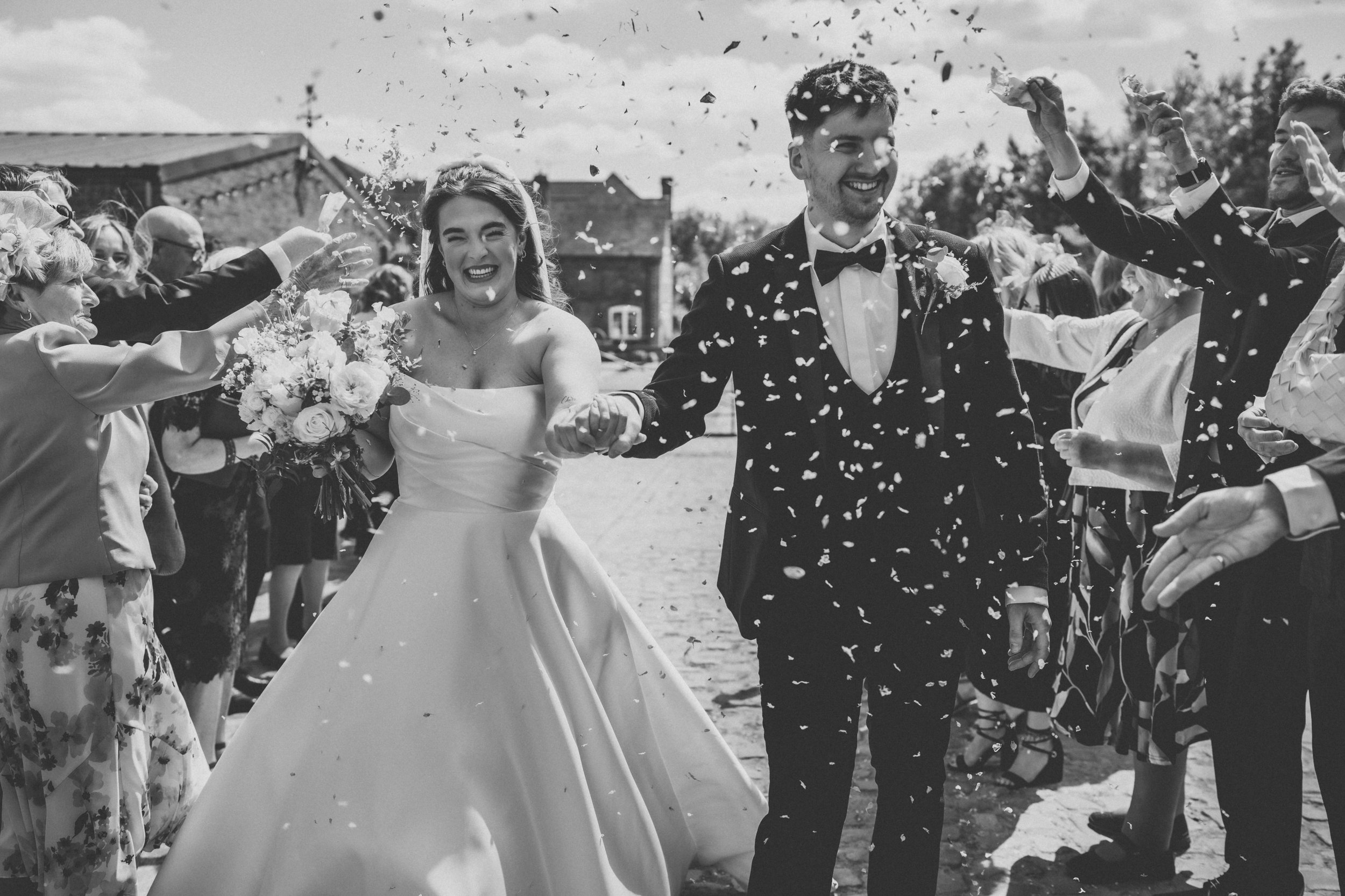 A black and white photo of a newlywed couple walking hand in hand, smiling, as they are showered with confetti by guests during their outdoor wedding celebration.