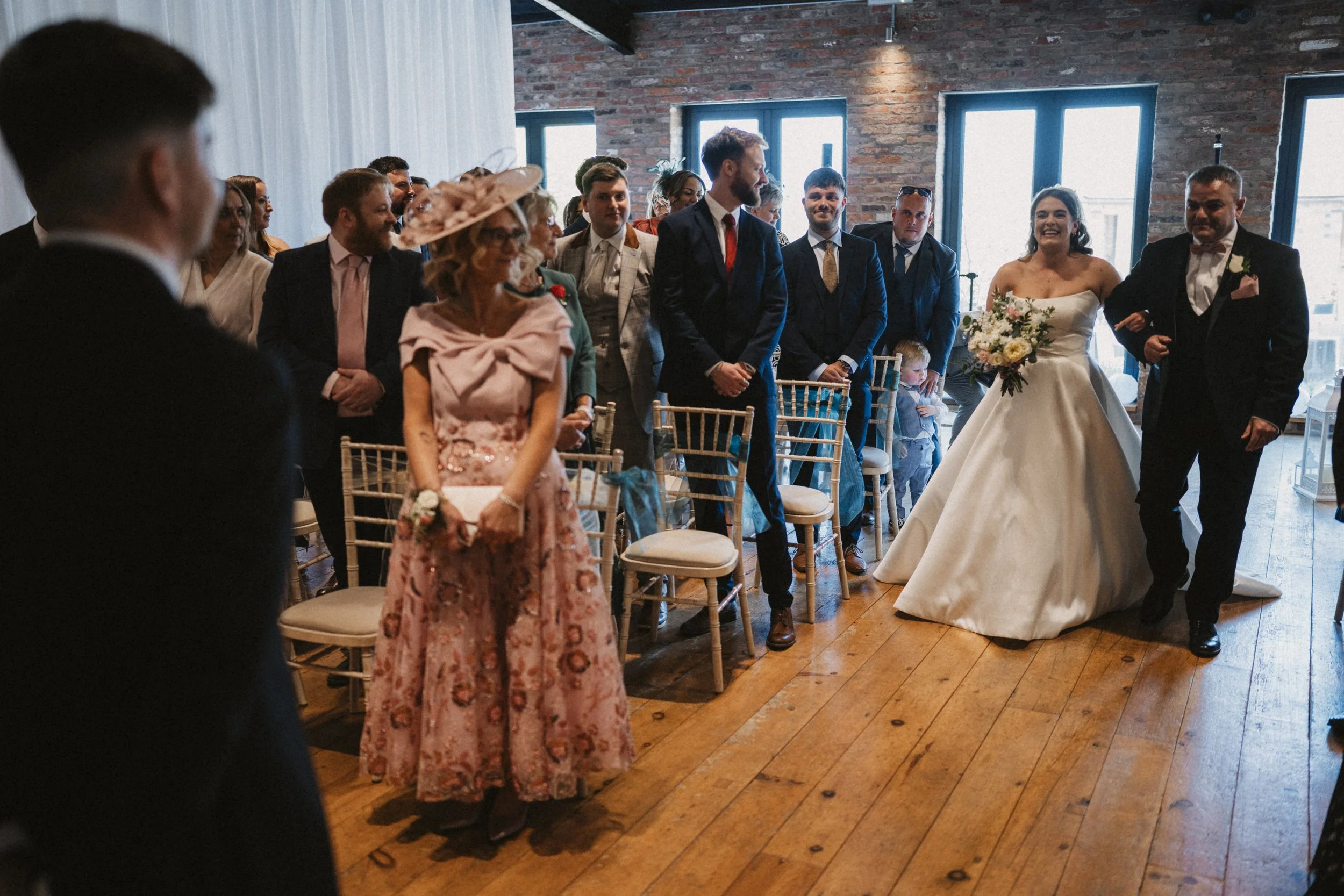 Bride and groom walking down aisle during wedding ceremony with guests standing and sitting, in a rustic venue with brick walls and large windows.