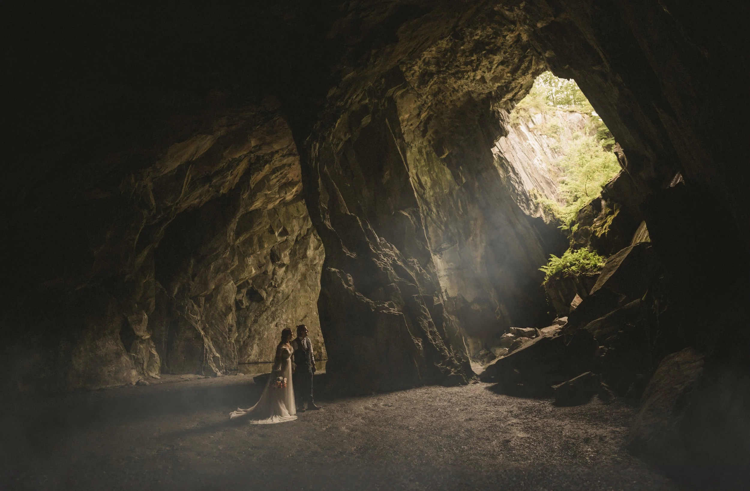 A couple in wedding attire stands inside a large cave with sunlight streaming in from the opening above, illuminating the rocky interior.