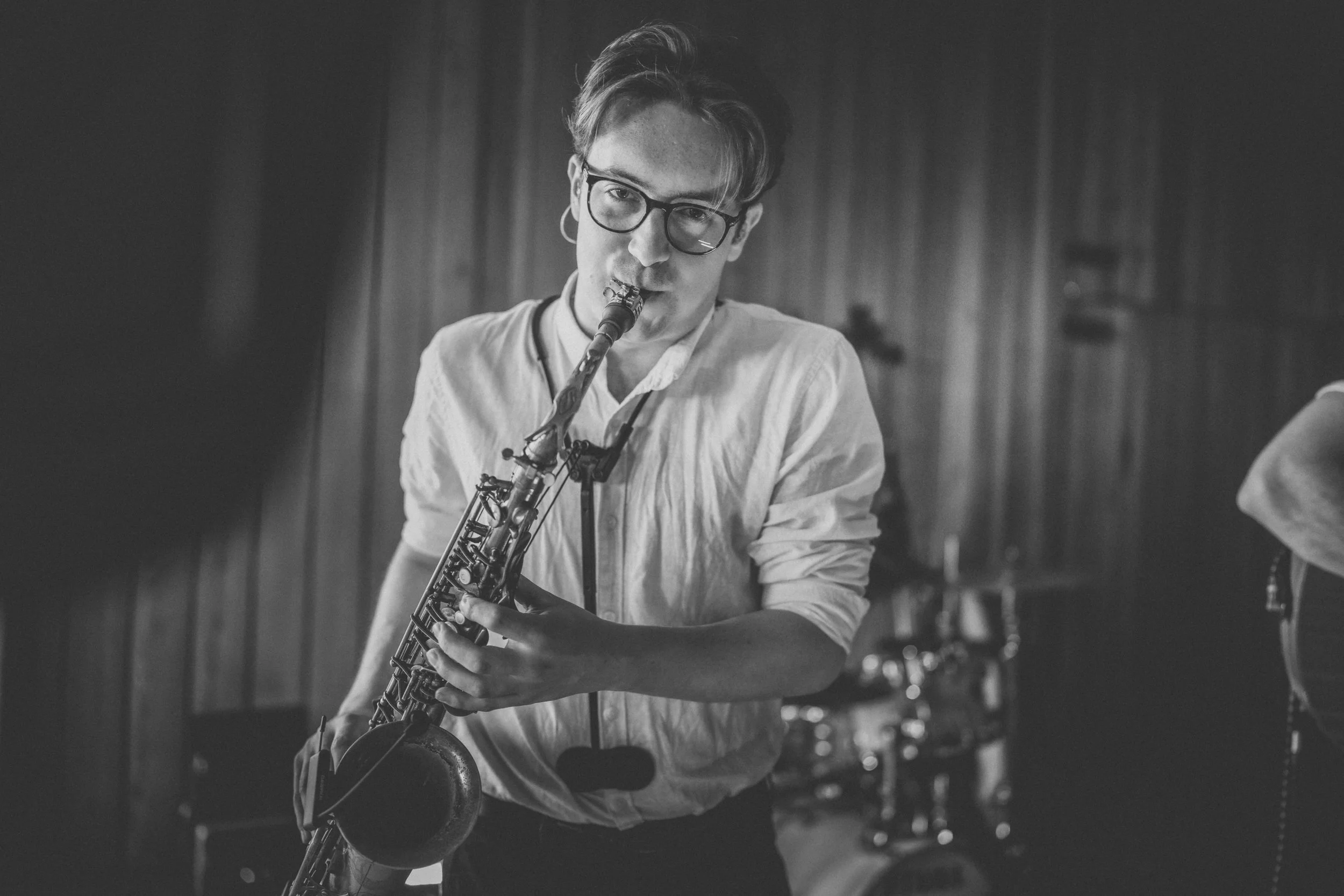 A young man with glasses and a white shirt playing the saxophone in a music studio.